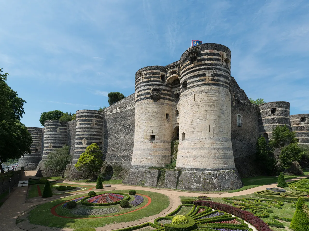 Les tours rayées de schiste sombre et de pierre claire du château d’Angers, au-dessus des parterres fleuris des jardins, sous un ciel bleu.