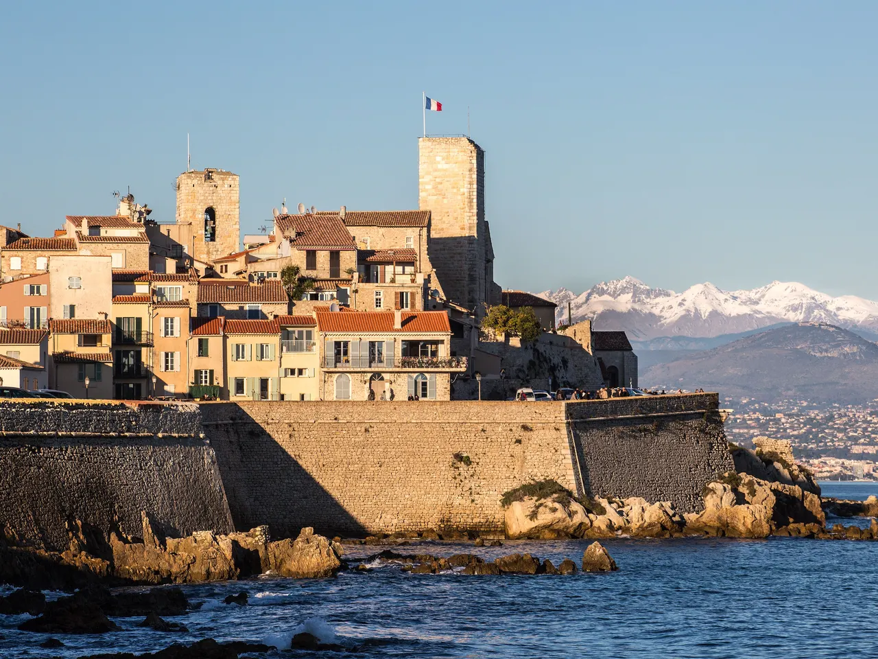 La vieille ville d’Antibes derrière ses remparts au bord de l’eau, dominée par les tours du château Grimaldi et de l’église, avec les Alpes enneigées au loin dans la lumière du soir.