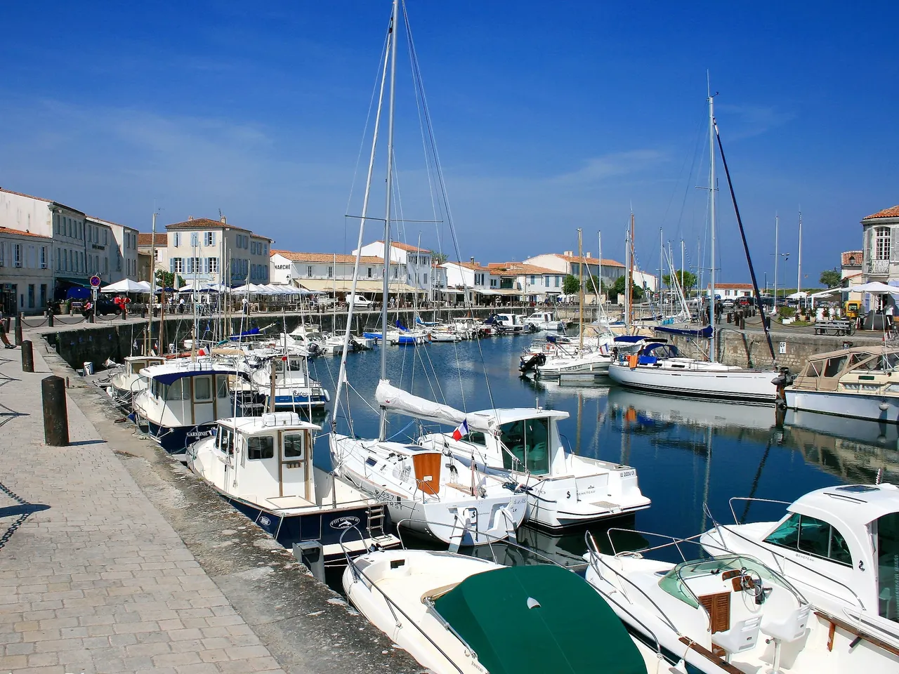 Le port d’Ars-en-Ré, son quai pavé longé de bateaux de plaisance et ses façades blanches au fond, sous un ciel d’un bleu profond.