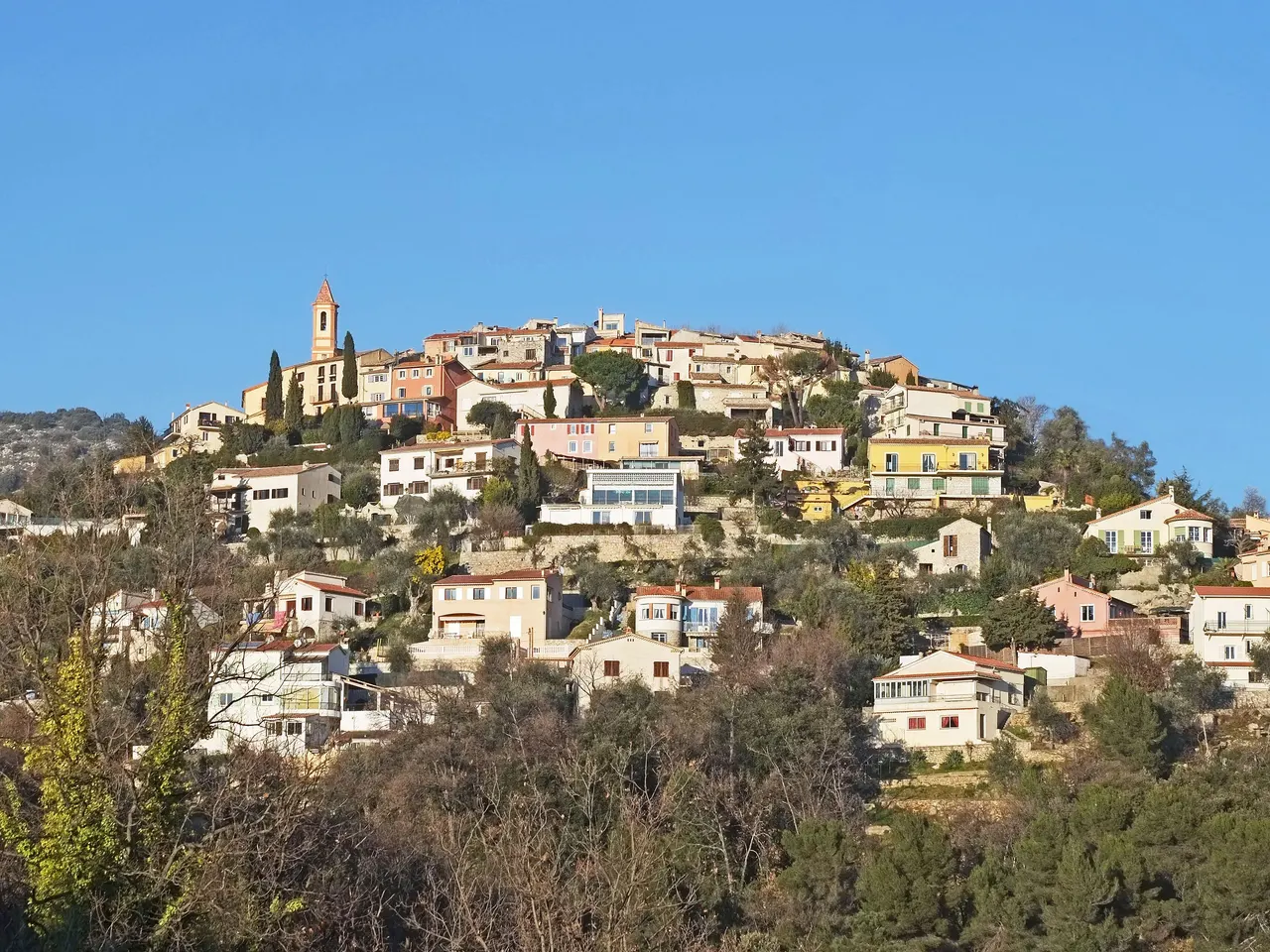 Le village d’Aspremont serré au sommet de sa colline, avec son clocher pointu et ses maisons colorées qui descendent la pente parmi les oliviers, sous un ciel bleu sans nuage.