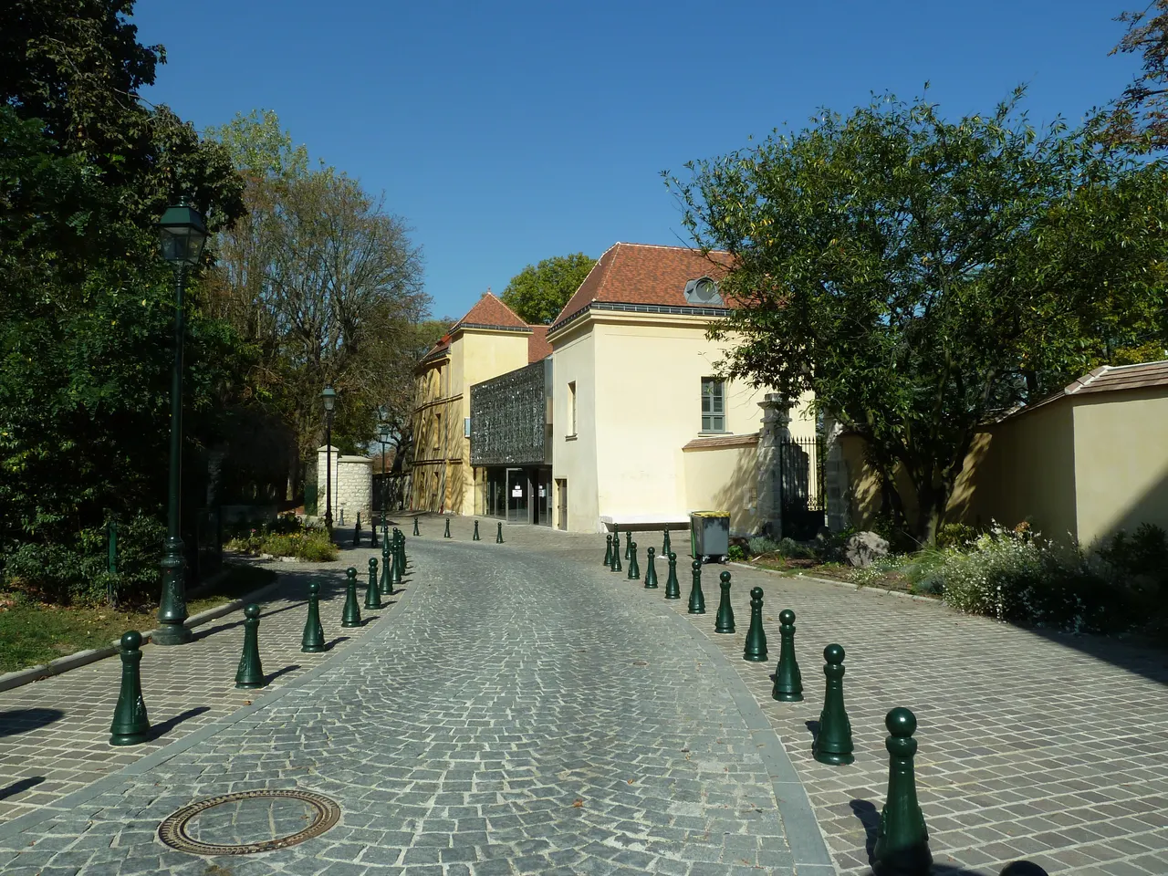 Une allée pavée bordée de bornes vertes mène à la maison de Richelieu, à Bagneux, bâtisse claire au toit de tuiles entre les arbres, sous un ciel bleu.
