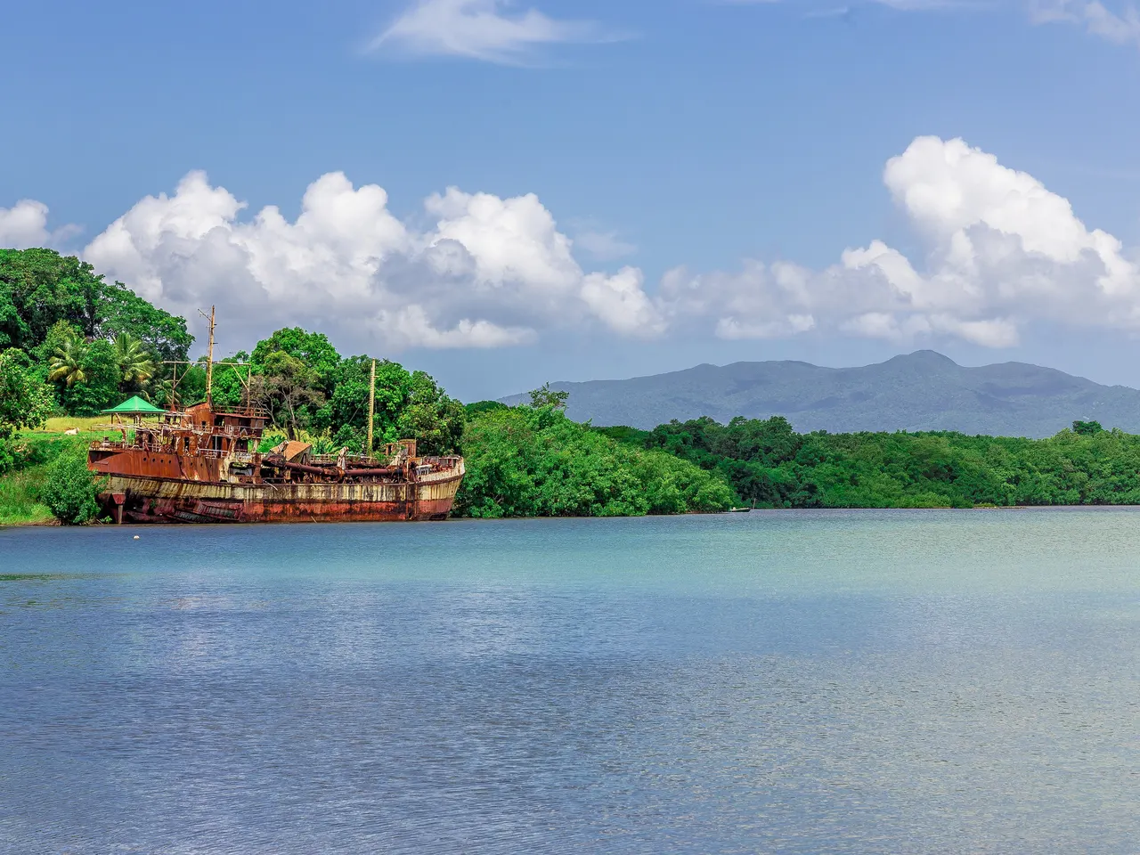 La baie de Baie-Mahault, avec l’épave rouillée du Bertina échouée contre la mangrove et des montagnes boisées à l’horizon, sous un ciel bleu.