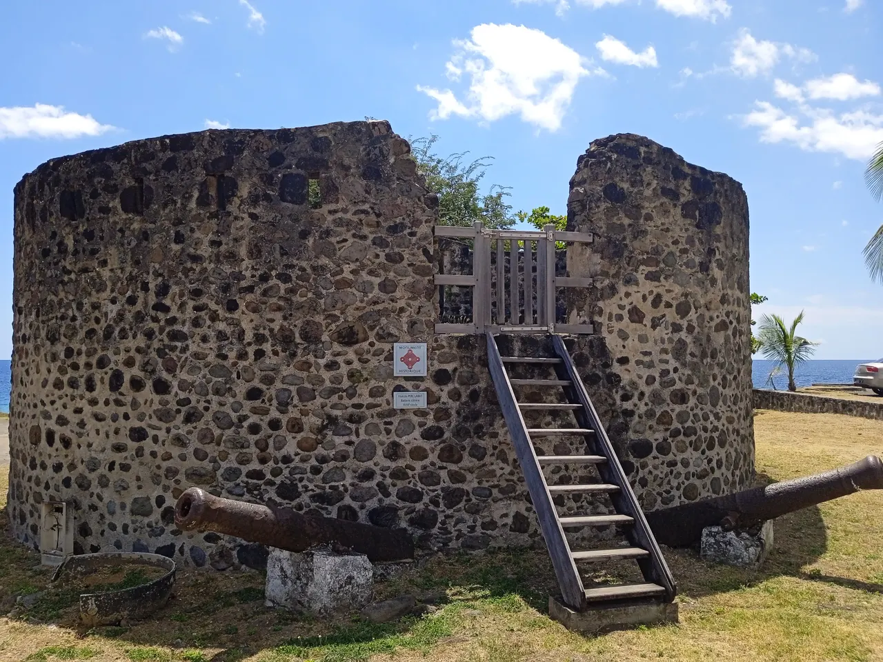 La tour du Père-Labat à Baillif : tour ronde en moellons de pierre, escalier de bois menant à une porte à claire-voie, deux canons anciens posés à son pied, la mer en arrière-plan.