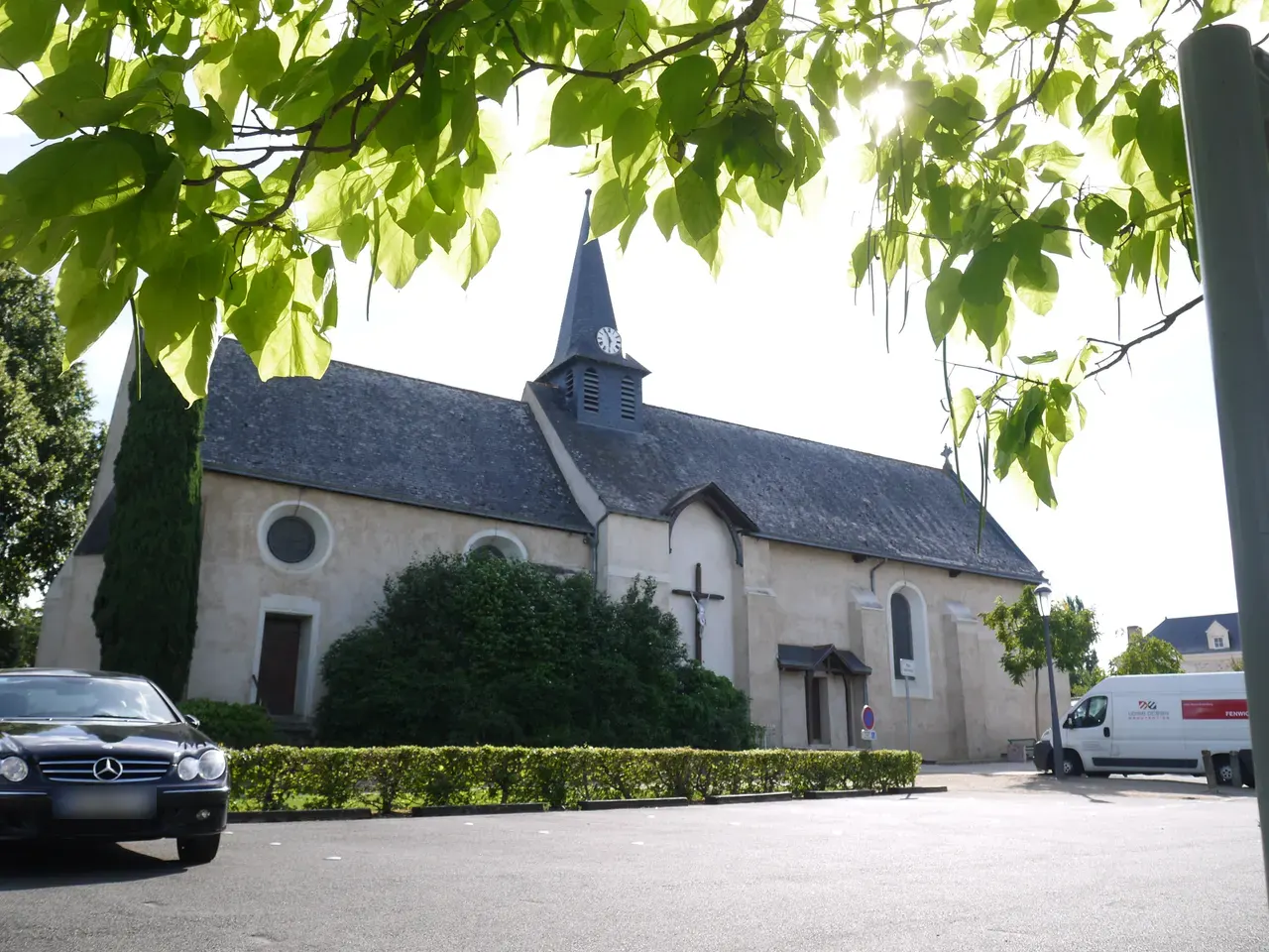 L’église Saint-Gilles de Beaucouzé, nef claire et clocher pointu en ardoise, vue sous un feuillage, avec une voiture et une camionnette garées devant.