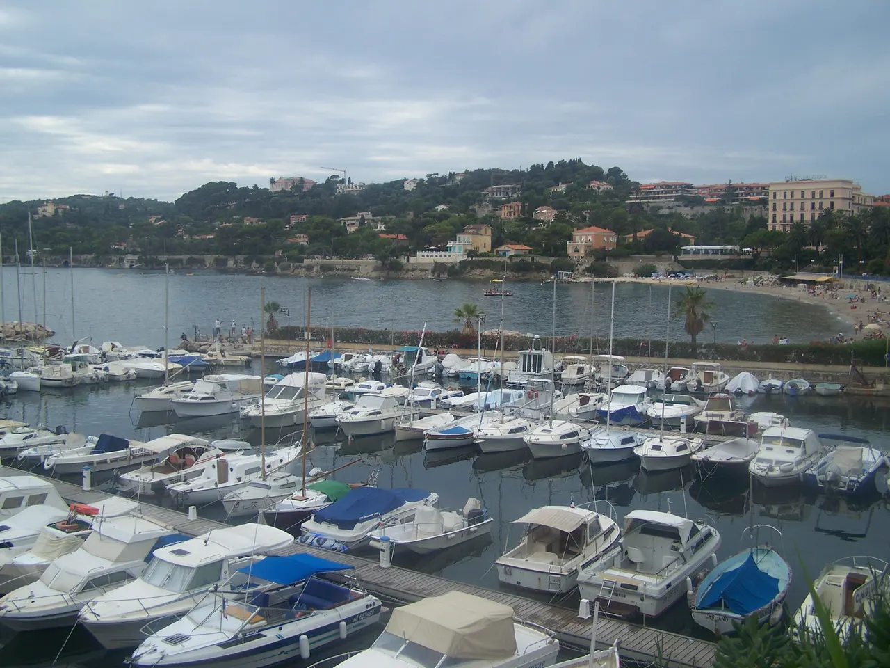 Le port de plaisance de Beaulieu-sur-Mer et ses bateaux, devant une plage et une colline boisée couverte de villas, sous un ciel nuageux.