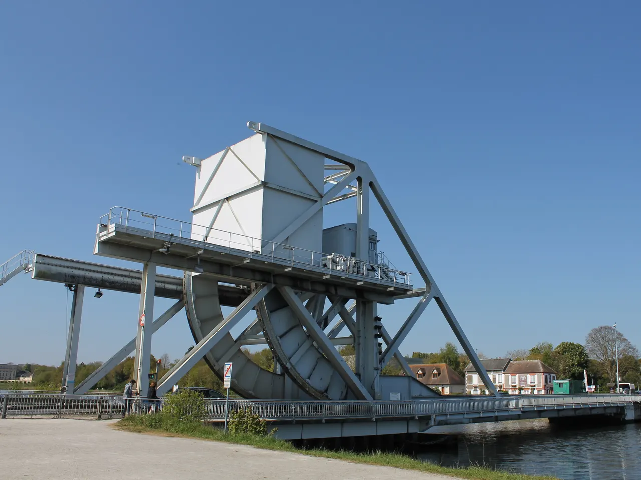 Pegasus Bridge à Bénouville, pont métallique gris clair surmonté d’un massif contrepoids et d’un grand quart de roue, au bord du canal de Caen à la mer, avec des maisons sur l’autre rive sous un ciel bleu