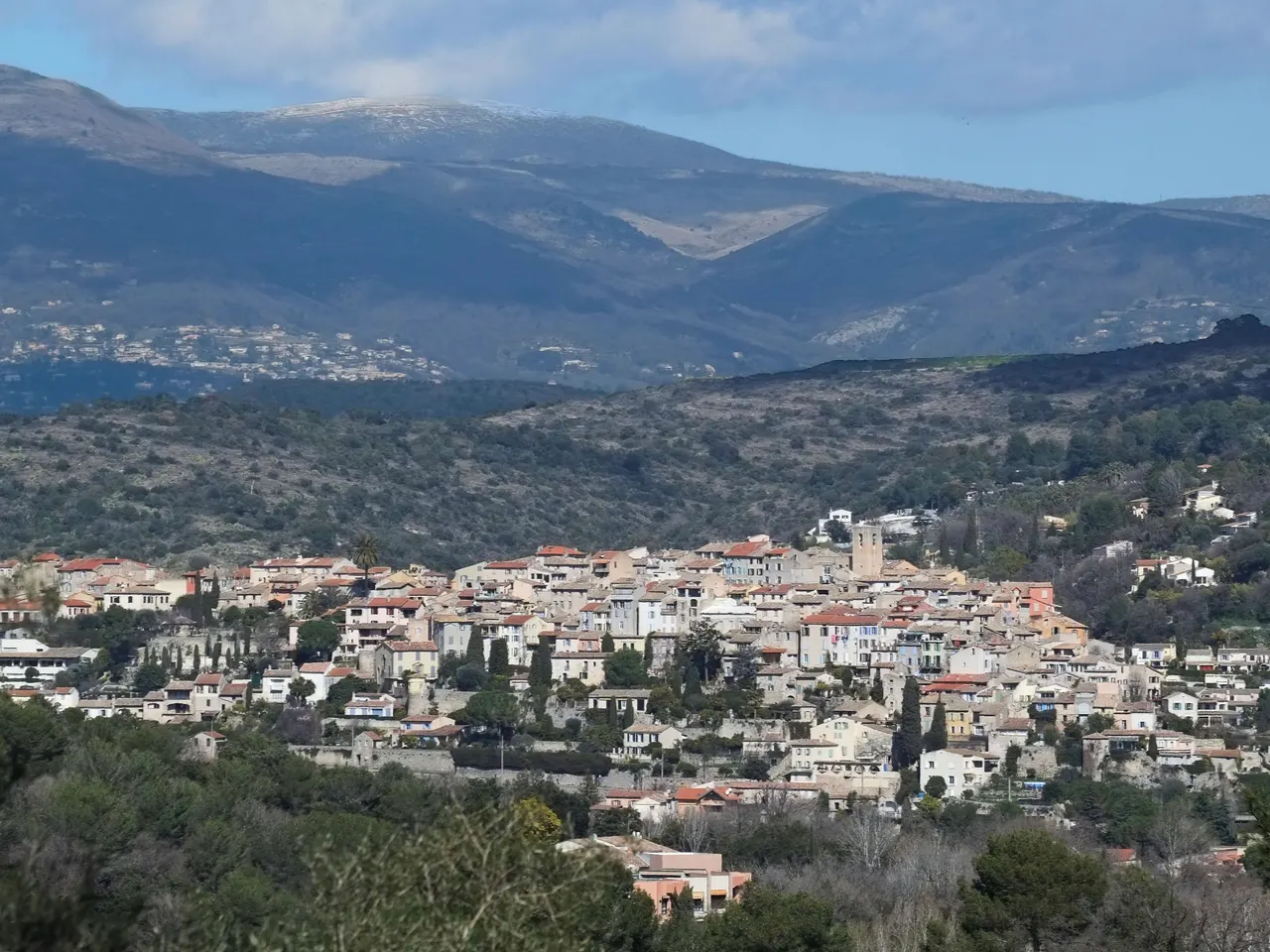 Le village perché de Biot et son clocher, serrés sur leur colline, devant des collines de garrigue et des montagnes en arrière-plan.