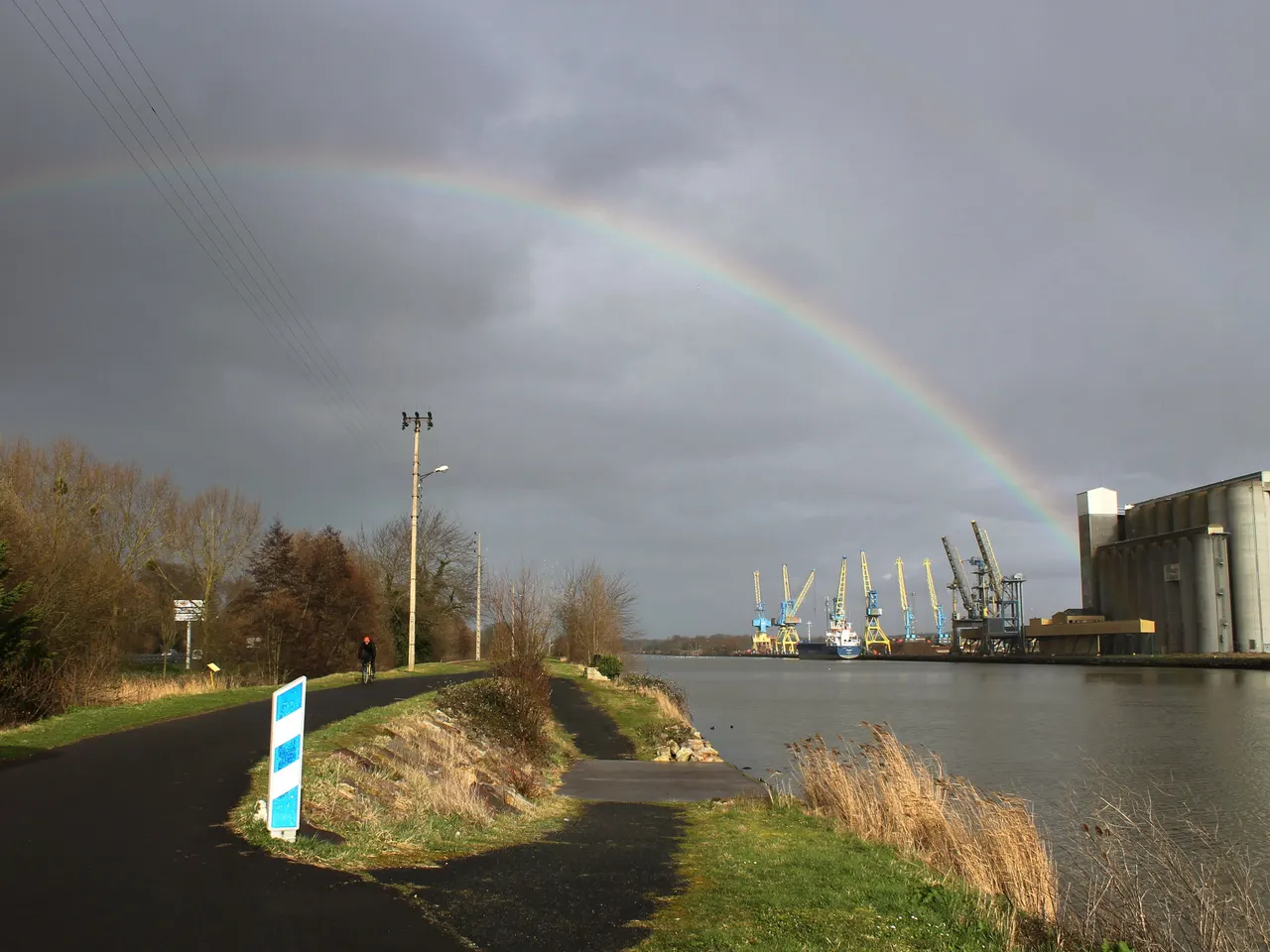 Le chemin de halage de Blainville-sur-Orne longeant le canal de Caen à la mer sous un ciel d’orage barré d’un arc-en-ciel, avec les grues du port et de grands silos sur l’autre rive
