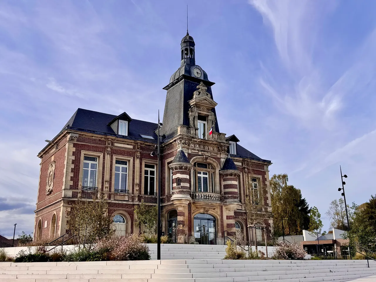 La mairie de Bois-Guillaume, en brique et pierre, avec son campanile à horloge, au-dessus d’un large escalier clair, dans la lumière de fin d’après-midi.