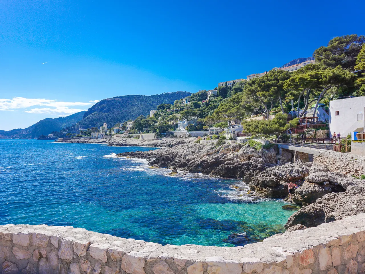 Le sentier du littoral de Cap-d’Ail longeant les rochers et une eau turquoise, avec des villas parmi les pins sur la colline et les montagnes de la côte au loin.