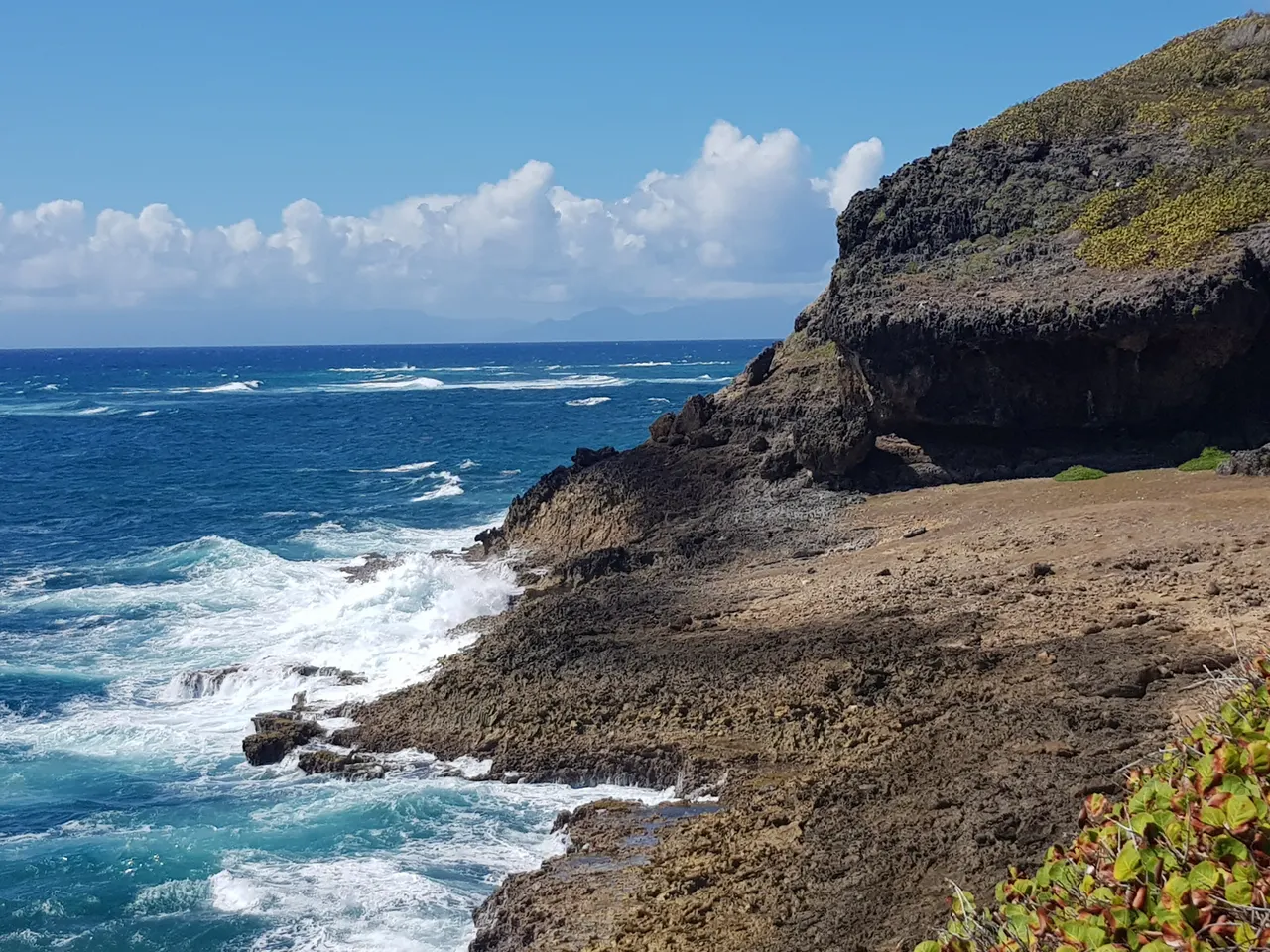 Les falaises des Galeries à Capesterre-de-Marie-Galante : une avancée de roche sombre en surplomb, un plateau rocheux et des vagues blanches qui frappent la côte sous un ciel clair
