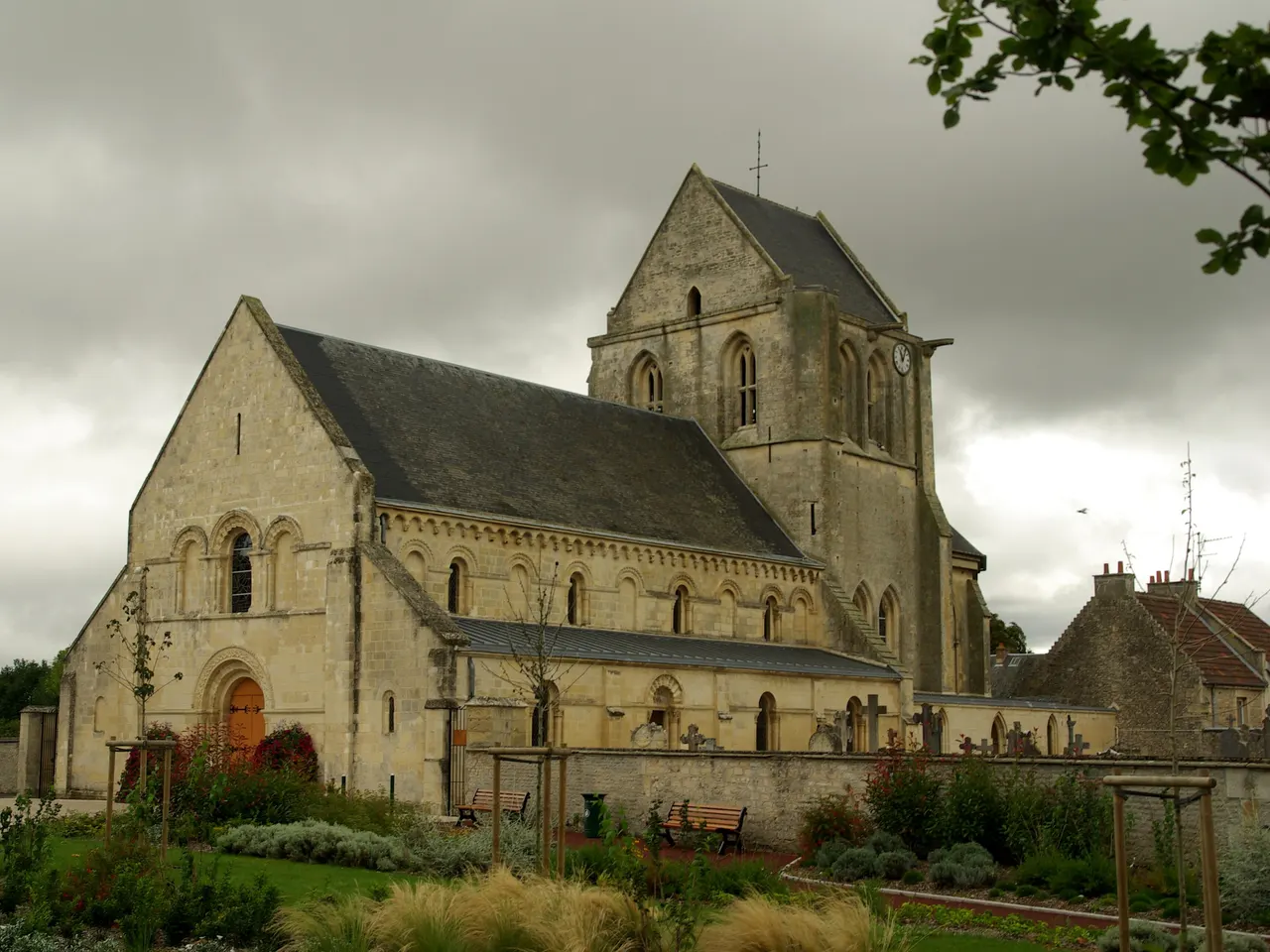 L’église Saint-Martin de Carpiquet, sa nef à arcatures romanes et son clocher, derrière un jardin planté, sous un ciel gris