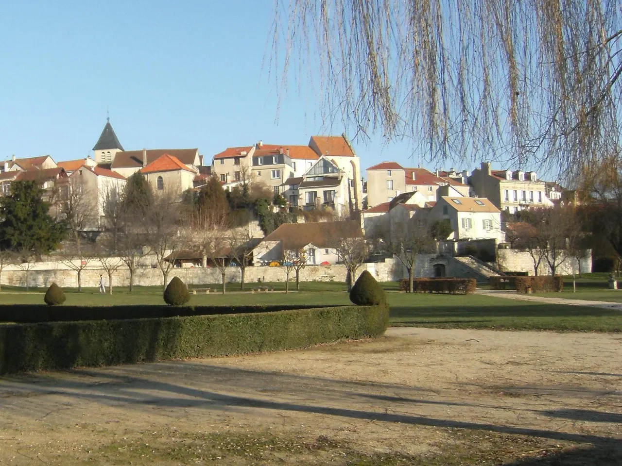 Le parc de la mairie de Carrières-sur-Seine, ses haies taillées et, au-dessus du mur de soutènement, les maisons du vieux village et le clocher de l’église.