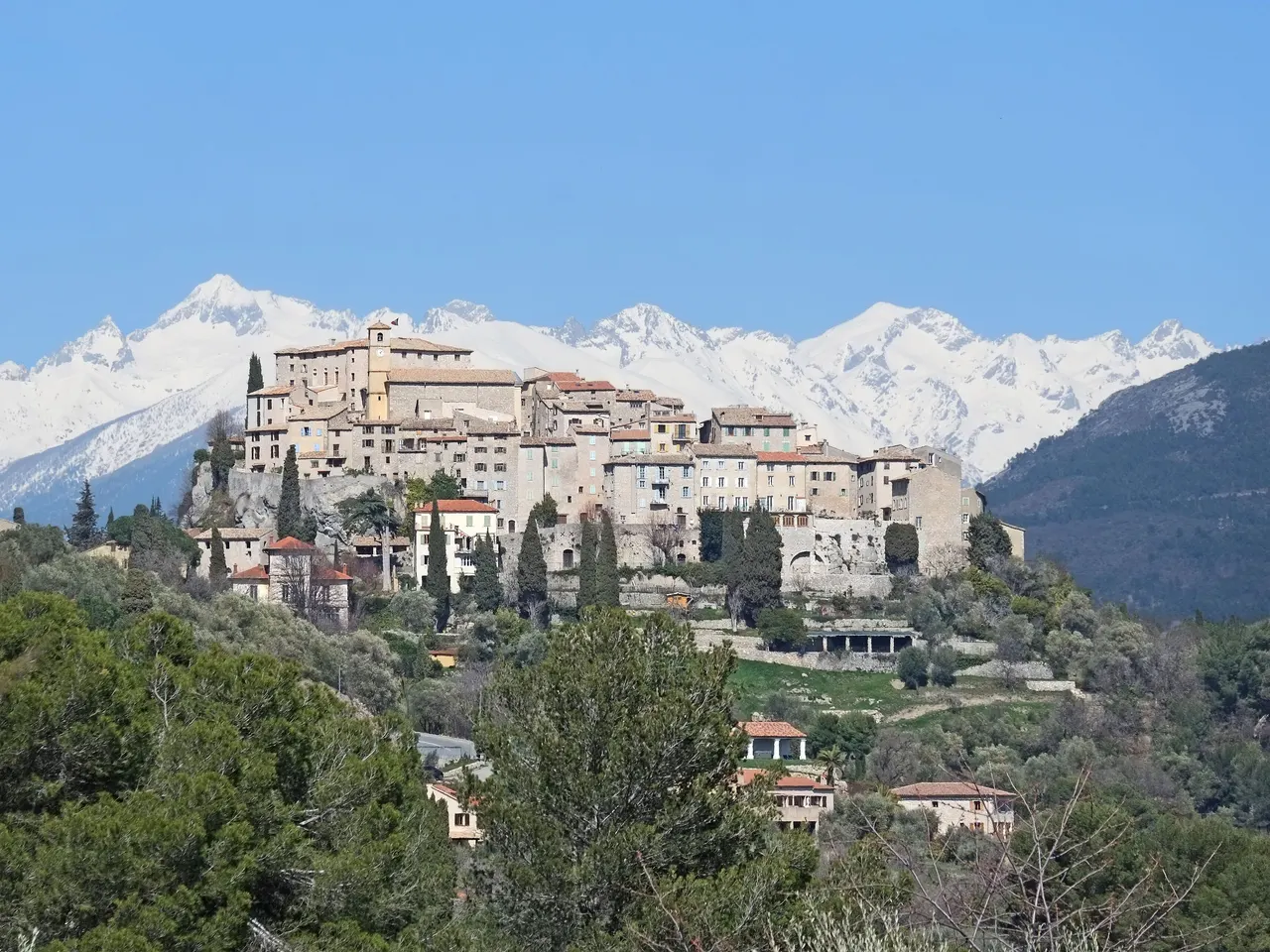 Le village perché de Carros, ses hautes maisons de pierre serrées sur leur colline, devant les sommets enneigés du Mercantour.
