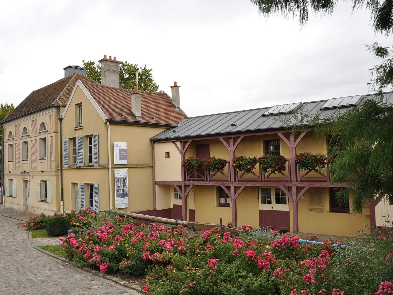 La maison Fournaise à Chatou, bâtiments jaunes à volets gris et galerie de bois fleurie, précédés d’un massif de roses