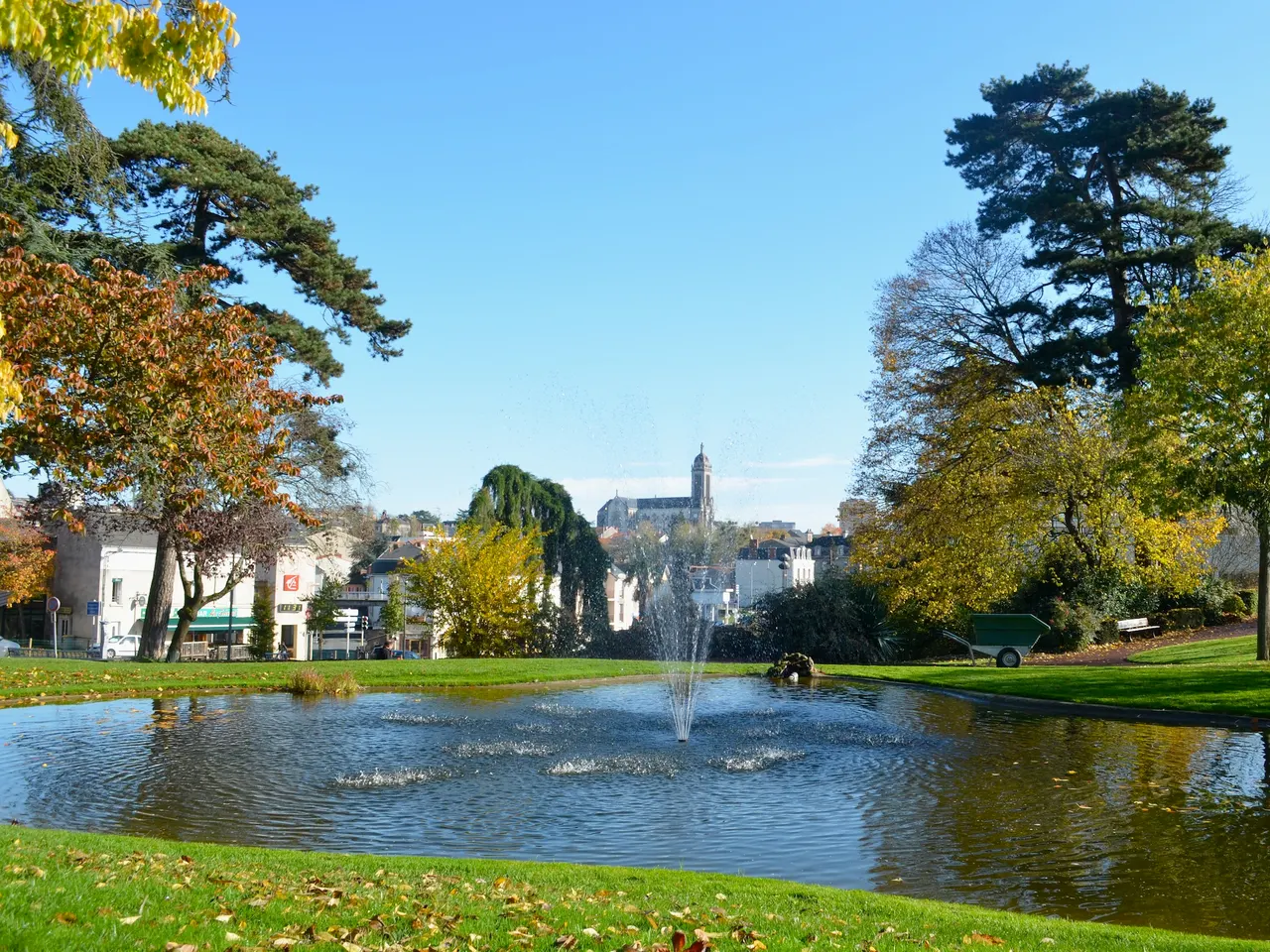 Le jardin du Mail à Cholet, bassin et jet d’eau au milieu des pelouses, entouré d’arbres aux couleurs d’automne, avec un clocher au loin.