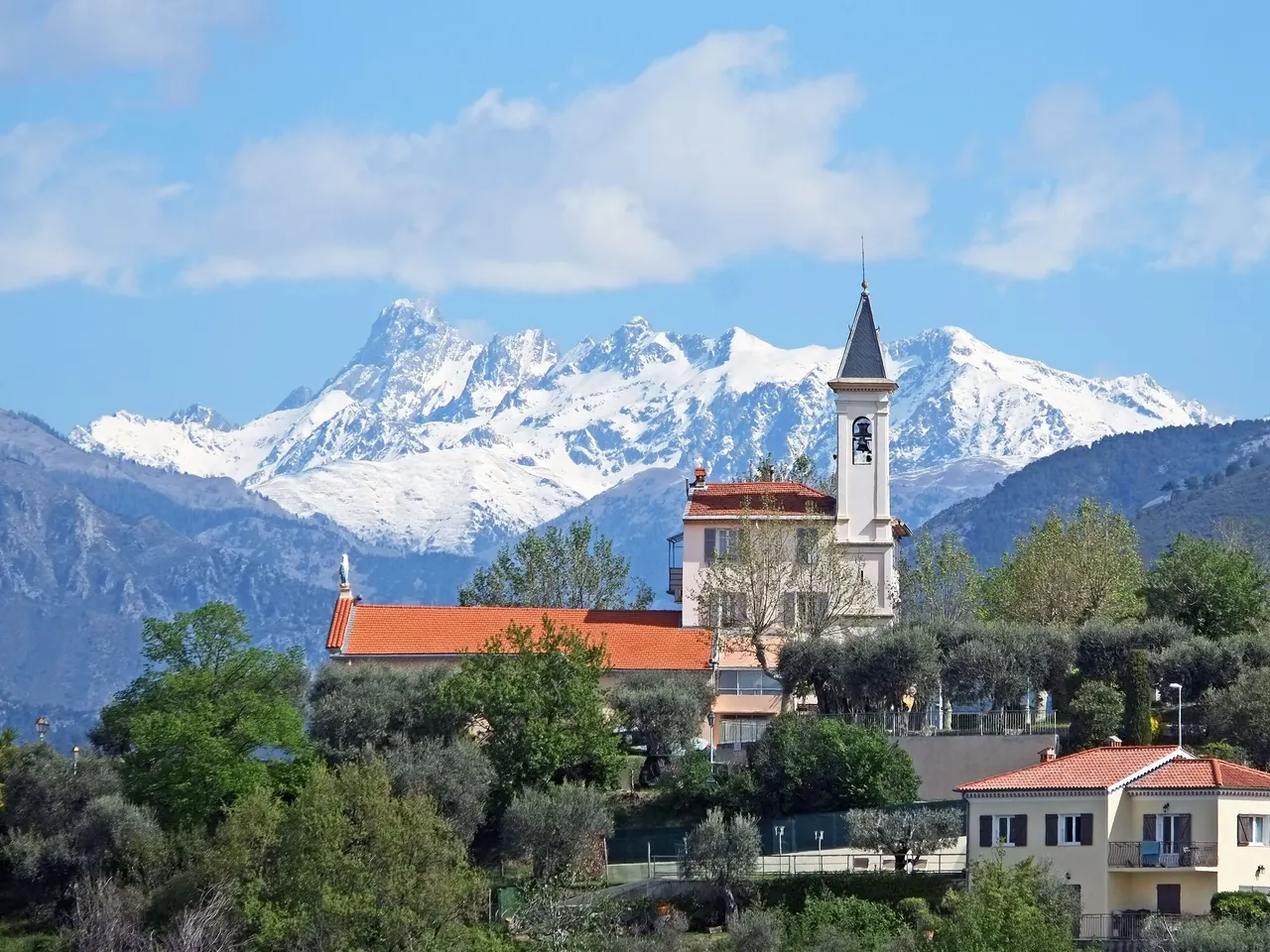 L’église Notre-Dame-de-la-Nativité de Colomars, son clocher clair et son toit de tuiles parmi les oliviers, devant les sommets enneigés des Alpes.