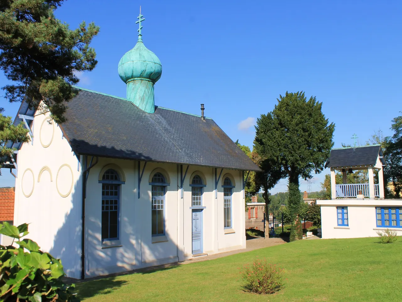 L’église orthodoxe Saint-Serge de Colombelles, petit édifice blanc aux fenêtres cintrées bleues coiffé d’un bulbe de cuivre vert-de-gris, et à droite sa cloche abritée sous un petit toit, au bord d’une pelouse et sous un ciel bleu