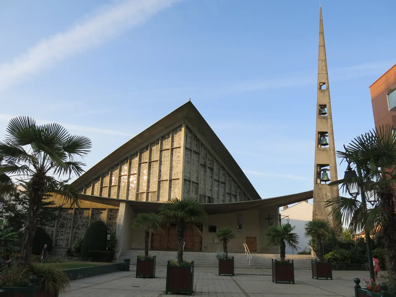 L’église Saint-Pierre-Saint-Paul de Colombes, son toit en pointe, sa façade ajourée et son clocher effilé, entre des palmiers en bacs
