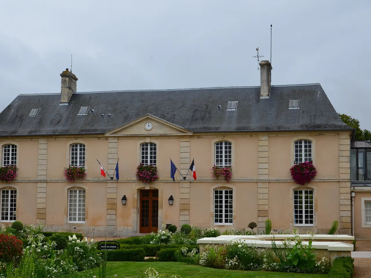 La façade classique de la mairie de Cormelles-le-Royal, fleurie aux fenêtres, derrière un jardin de massifs blancs