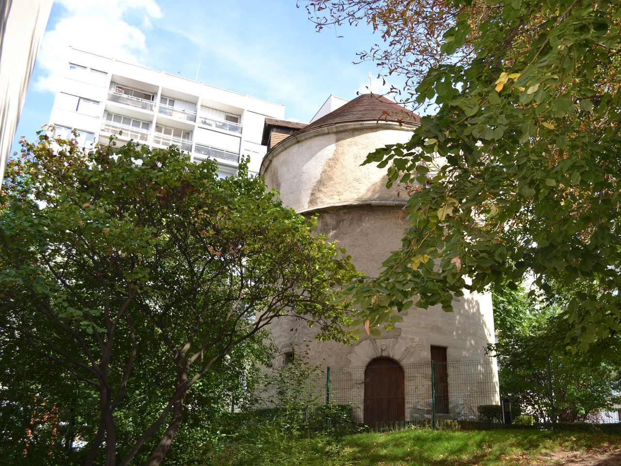 Le vieux colombier de Créteil, tour ronde en pierre coiffée d’un toit conique, entre les arbres, devant un immeuble blanc.