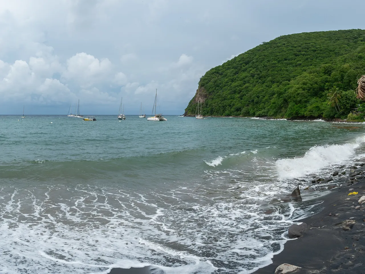 L’anse de Deshaies sous un ciel chargé : voiliers au mouillage, vagues sur une grève de sable noir et de galets, et un morne couvert de forêt qui ferme la baie.