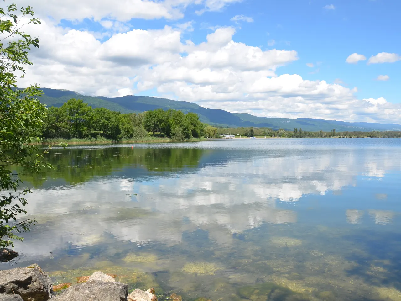 Le lac de Divonne-les-Bains, dont l’eau claire reflète les nuages, bordé d’arbres au pied de la chaîne du Jura.