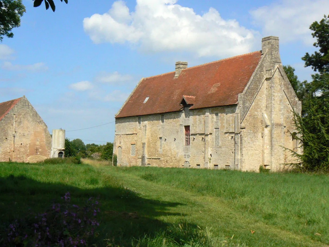 Le manoir de la Baronnie à Douvres-la-Délivrande, long bâtiment médiéval en pierre claire au toit de tuiles rouges, au fond d’une prairie d’herbe haute, avec un second bâtiment de pierre plus petit à gauche
