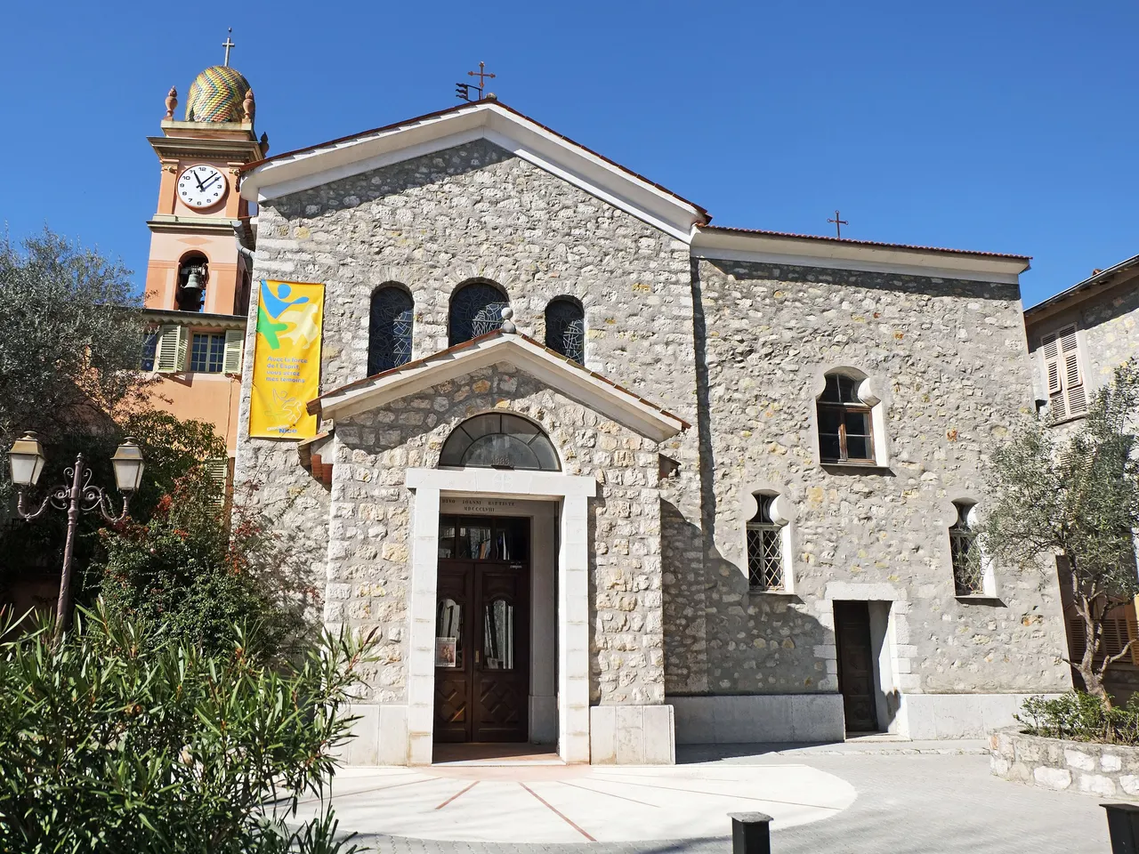 La façade en pierre de l’église Saint-Jean-Baptiste de Drap, avec son clocher orangé à horloge surmonté d’un dôme de tuiles colorées, sous un ciel bleu.