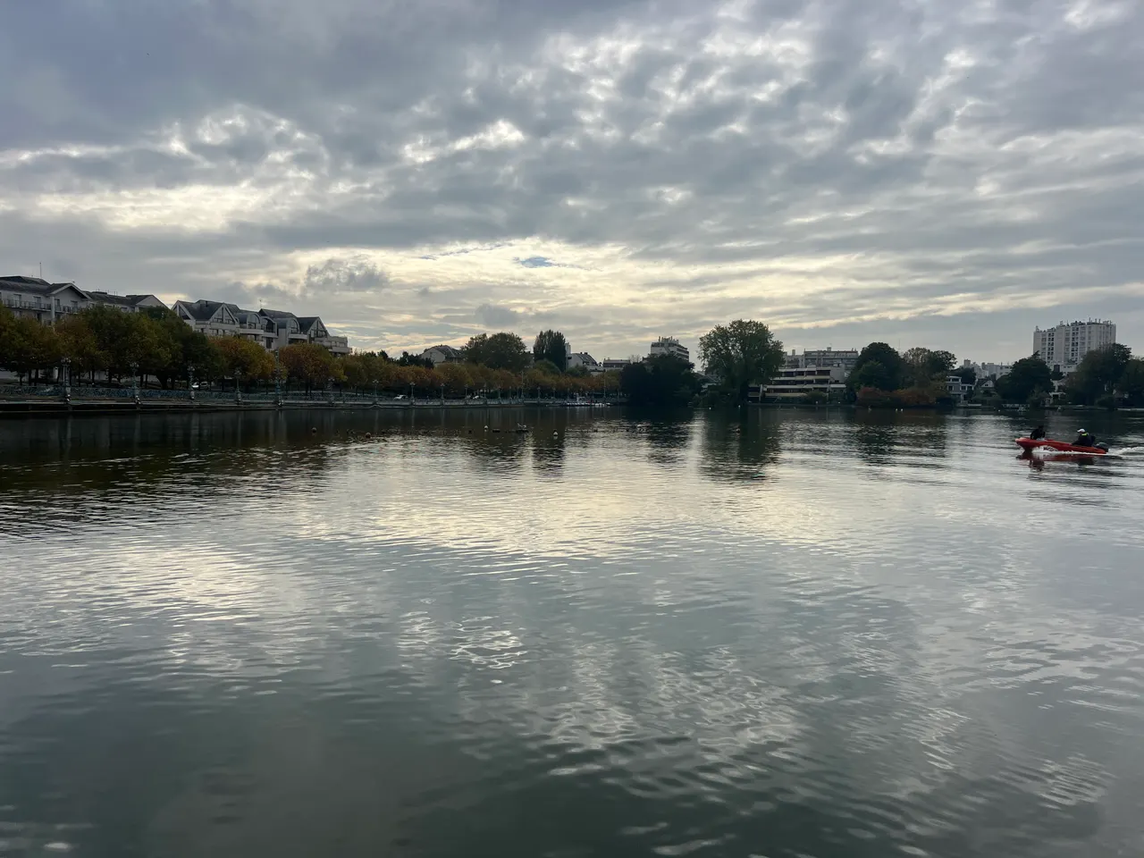 Le lac d’Enghien sous un ciel nuageux : rive bordée d’arbres d’automne et d’immeubles, une petite embarcation rouge filant sur l’eau.