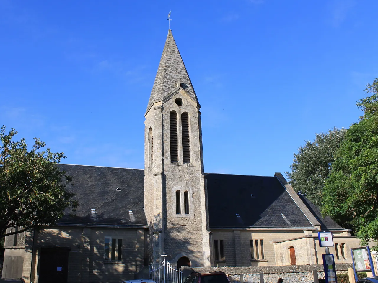 L’église Saint-Ursin d’Épron, son clocher de pierre à flèche pointue entre deux ailes aux toits d’ardoise, encadrée d’arbres sous un ciel d’un bleu intense