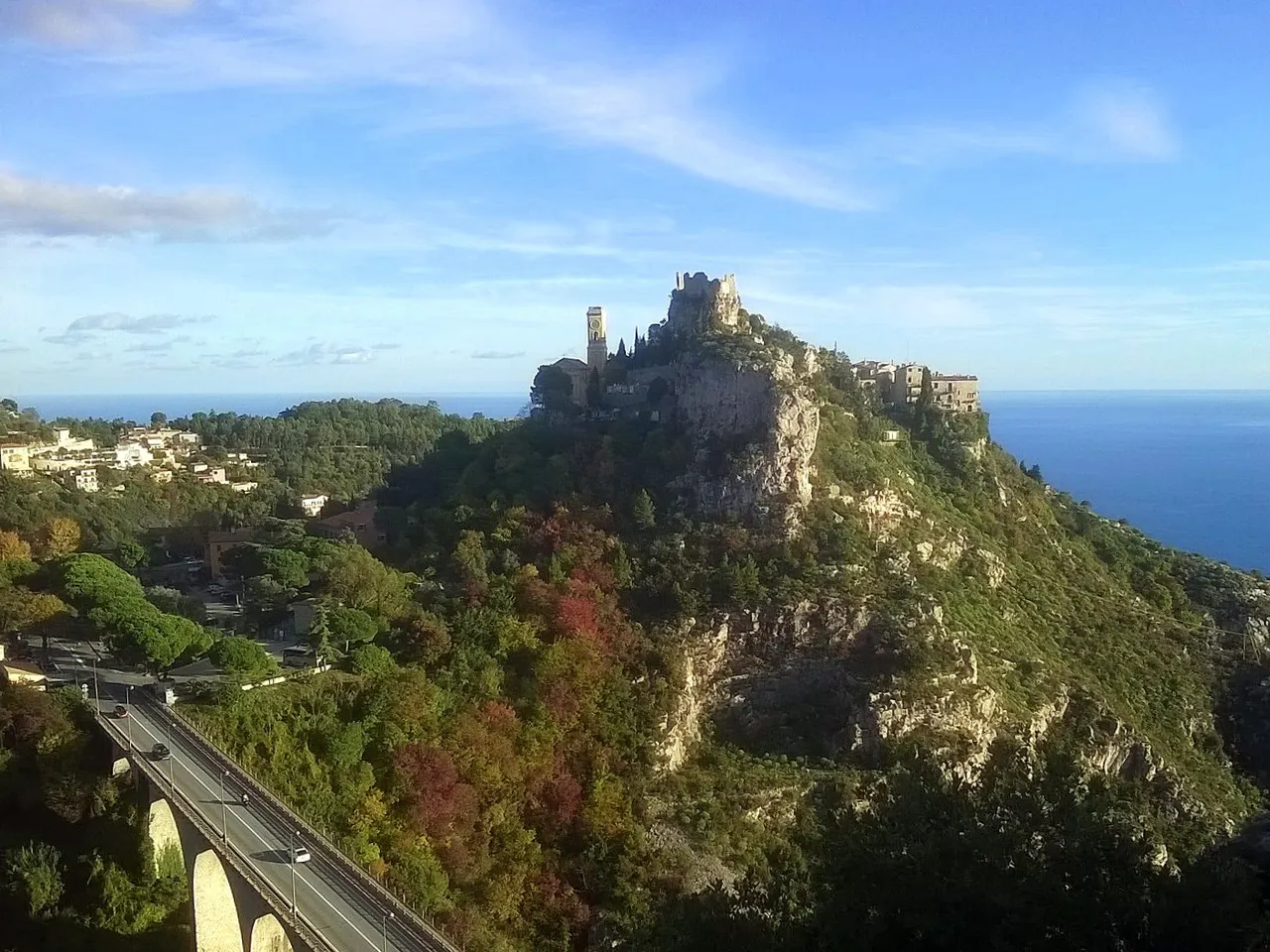 Le village perché d’Èze sur son piton rocheux, avec les ruines du château et le clocher de l’église au sommet, la mer à l’horizon et un viaduc routier au premier plan.