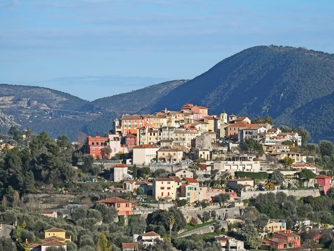 Le village perché de Falicon, ses maisons ocre, roses et jaunes étagées sur la colline autour d’un clocher, devant des montagnes boisées sous un ciel bleu.