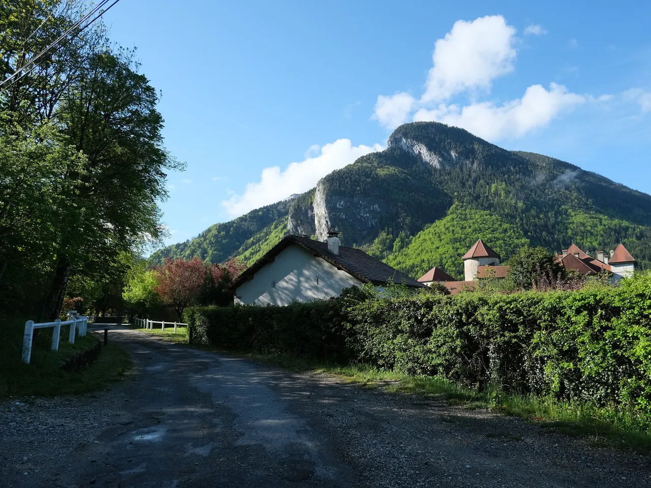 Un chemin de campagne à Thorens-Glières, dans la commune de Fillière : haies, maison blanche et tours aux toits de tuiles du château de Thorens au pied d’une montagne boisée.