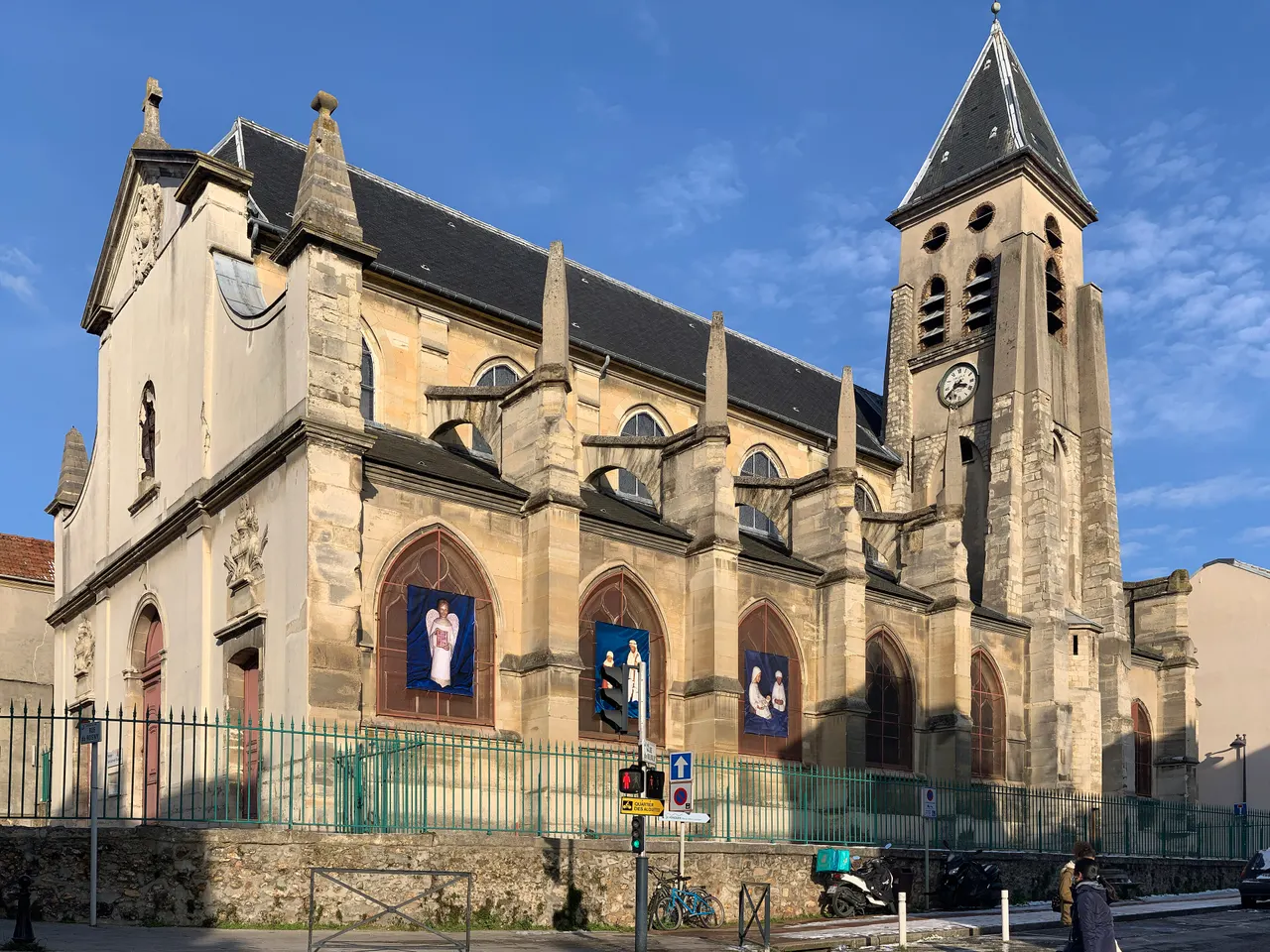 L’église Saint-Germain-l’Auxerrois de Fontenay-sous-Bois, sa nef épaulée d’arcs-boutants et son clocher à horloge, derrière une grille verte, sous un ciel bleu.