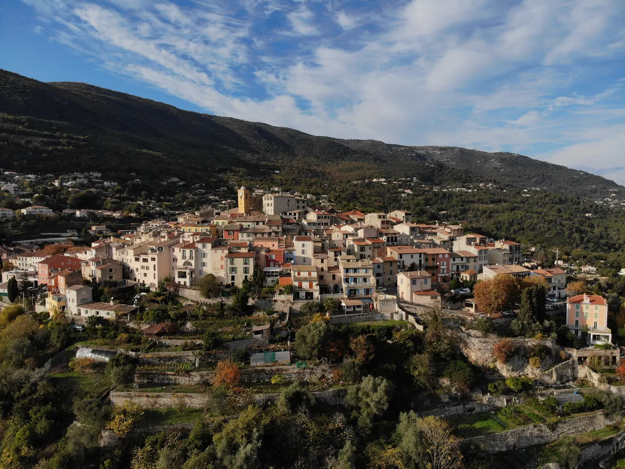 Vue aérienne de Gattières : des maisons colorées serrées autour d’un clocher ocre, au-dessus de jardins en terrasses, sur fond de collines boisées.