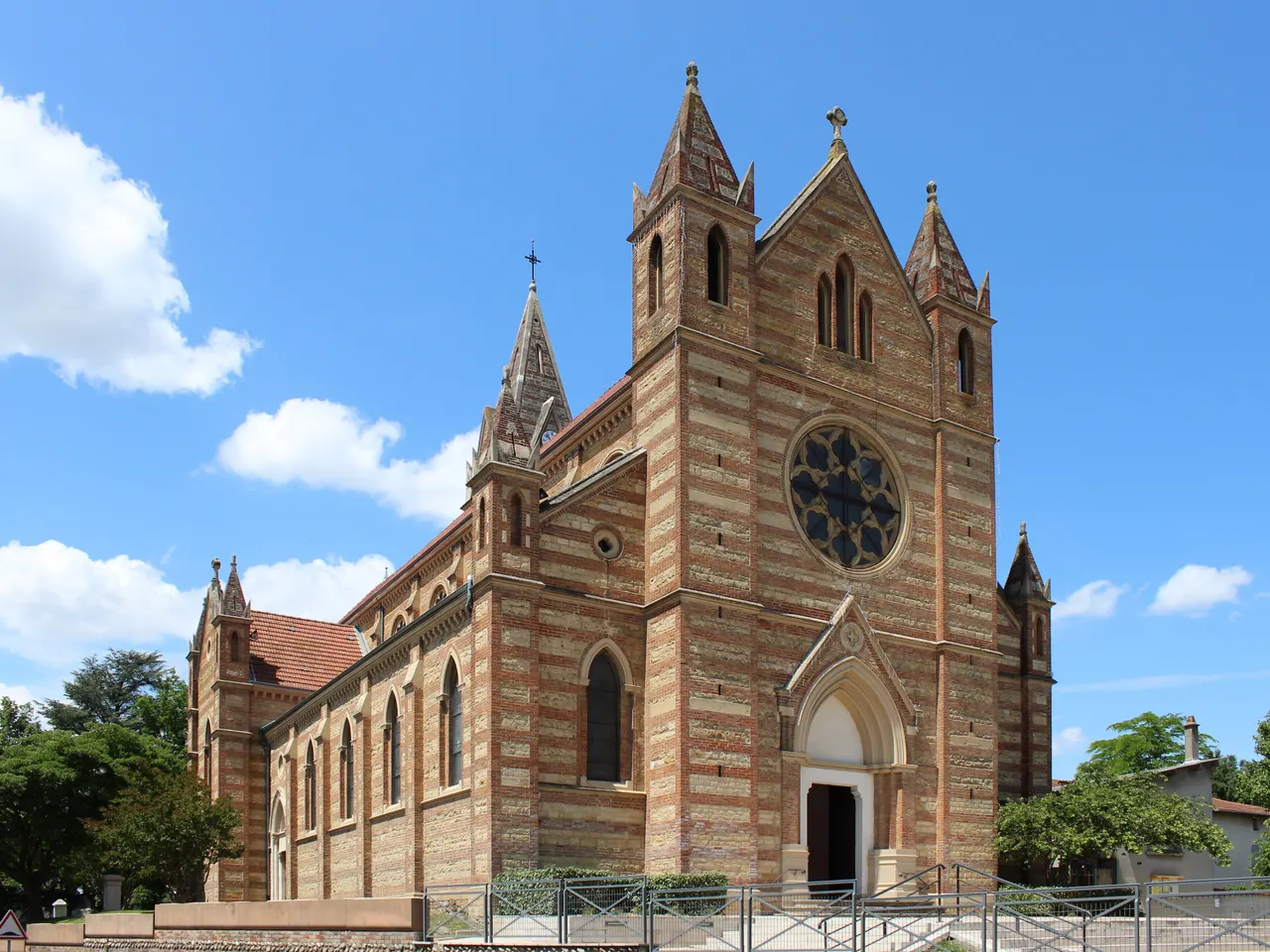 L’église Saint-Barthélemy de Genas, de style néogothique, avec ses assises alternées de pierre et de brique, sa rosace et ses clochetons, sous un ciel bleu.