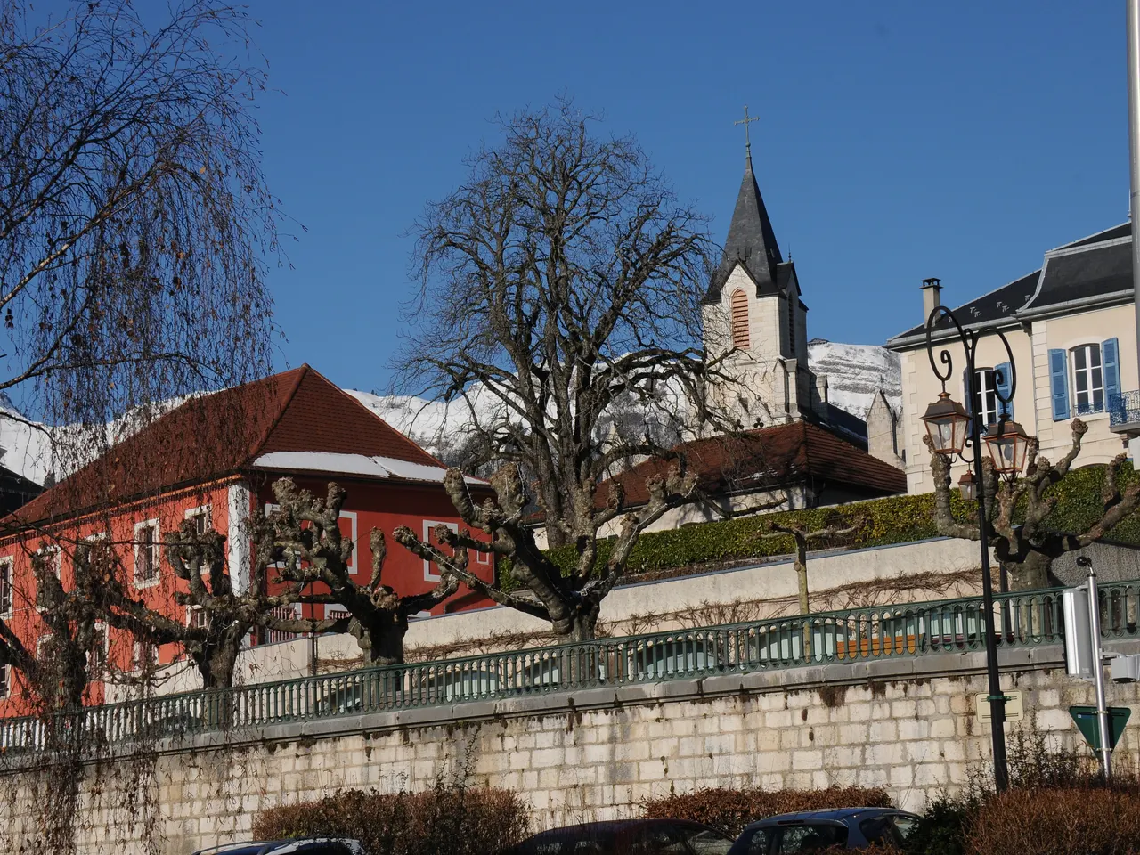 Le clocher de l’église de Gex dépassant d’une maison rouge et d’arbres taillés, devant les sommets enneigés du Jura, sous un ciel bleu.
