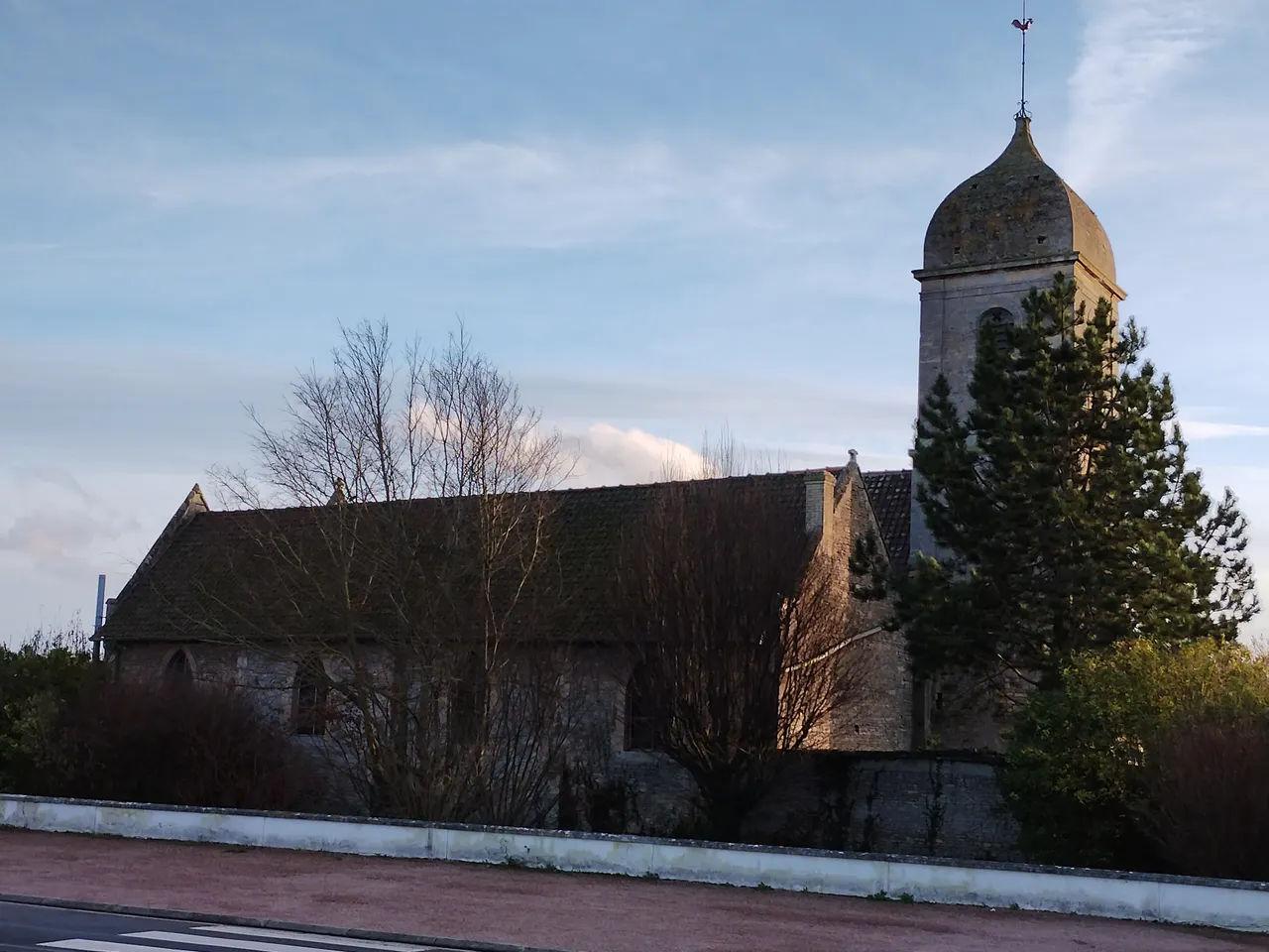 L’église Saint-Martin de Giberville, sa longue nef de pierre et son clocher coiffé d’un dôme à girouette, derrière des arbres dénudés et un muret blanc, dans la lumière rasante d’une fin de journée