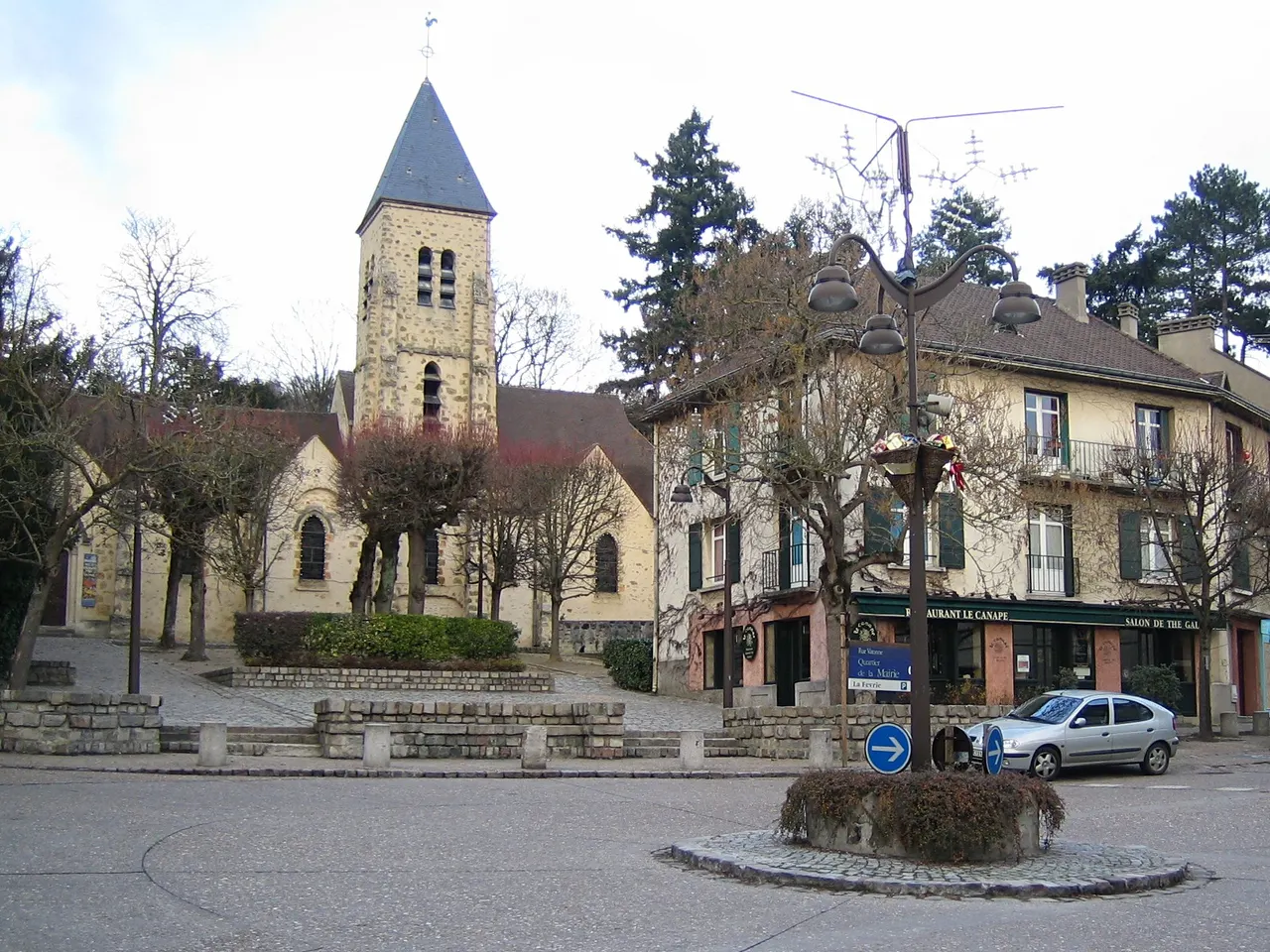 La place de l’église de Gif-sur-Yvette, le clocher de pierre de Saint-Rémi et une maison à balcons autour d’un petit rond-point, sous un ciel couvert.