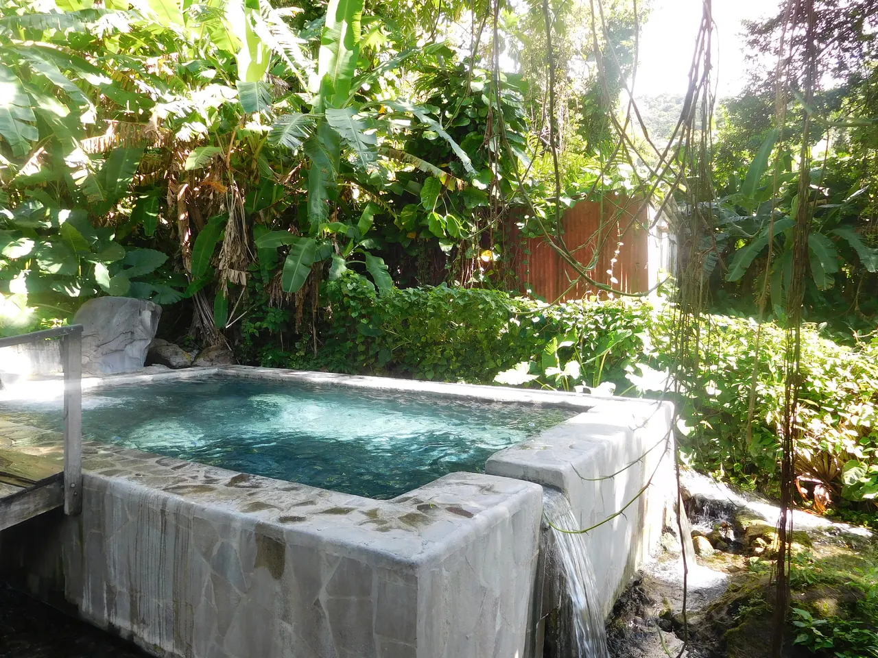 Un bassin des sources chaudes de Dolé, eau turquoise retenue par un muret de pierre, au milieu d’une végétation tropicale dense et ensoleillée.