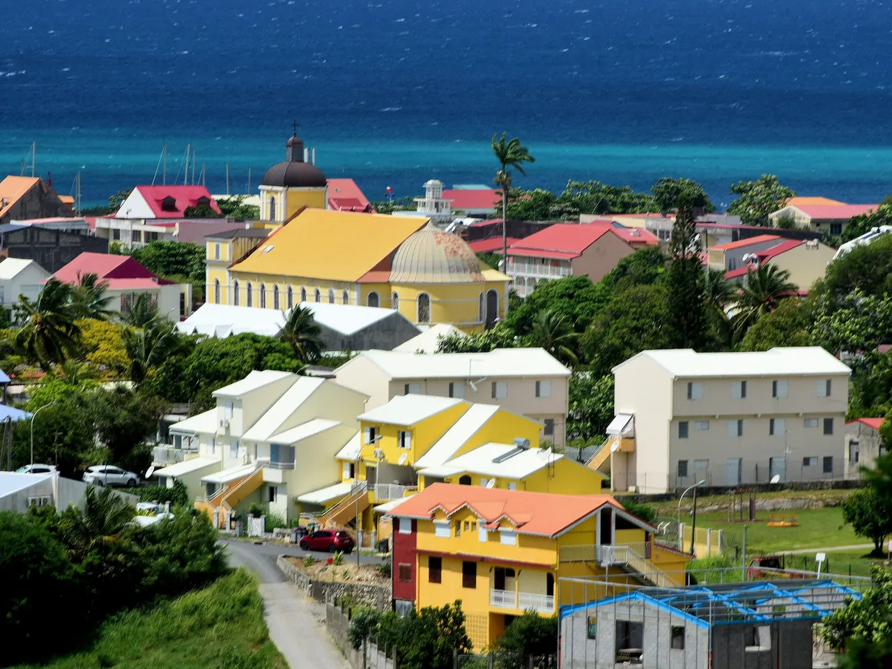 Grand-Bourg vu des hauteurs : l’église de l’Immaculée-Conception, jaune à coupole, entourée de maisons colorées aux toits rouges, devant une mer d’un bleu profond