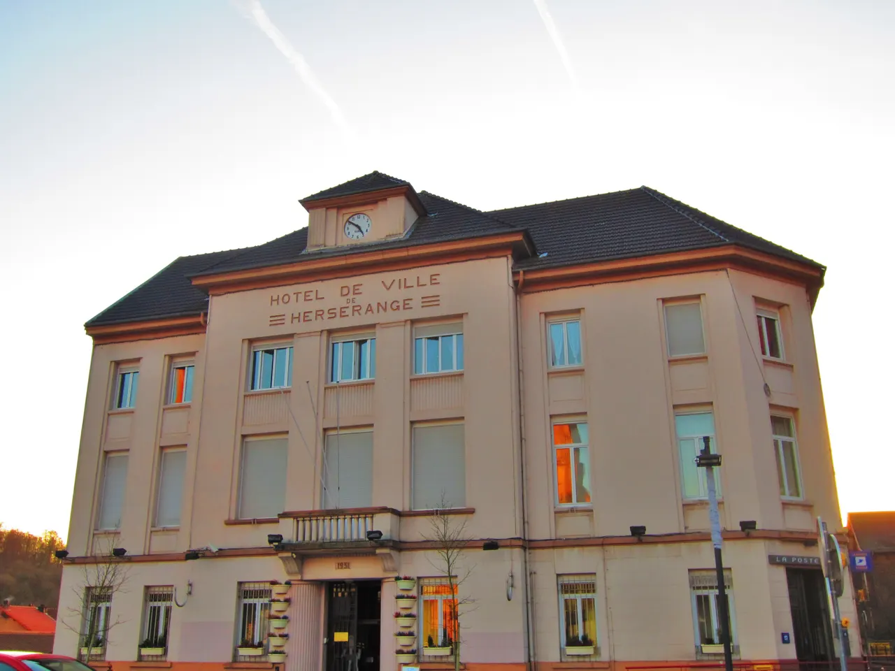 Façade de l’hôtel de ville de Herserange, bâtiment clair surmonté d’une horloge, éclairé par le soleil couchant.