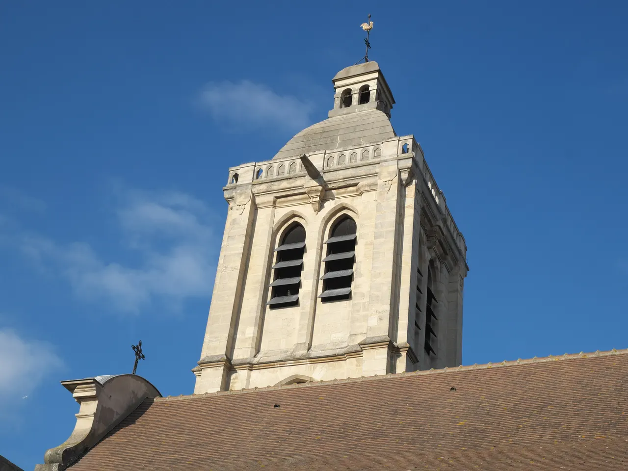 Le clocher de pierre claire de l’église Saint-Nicolas de Houilles, coiffé d’un lanternon et d’un coq, au-dessus du toit de tuiles, sous un ciel bleu.