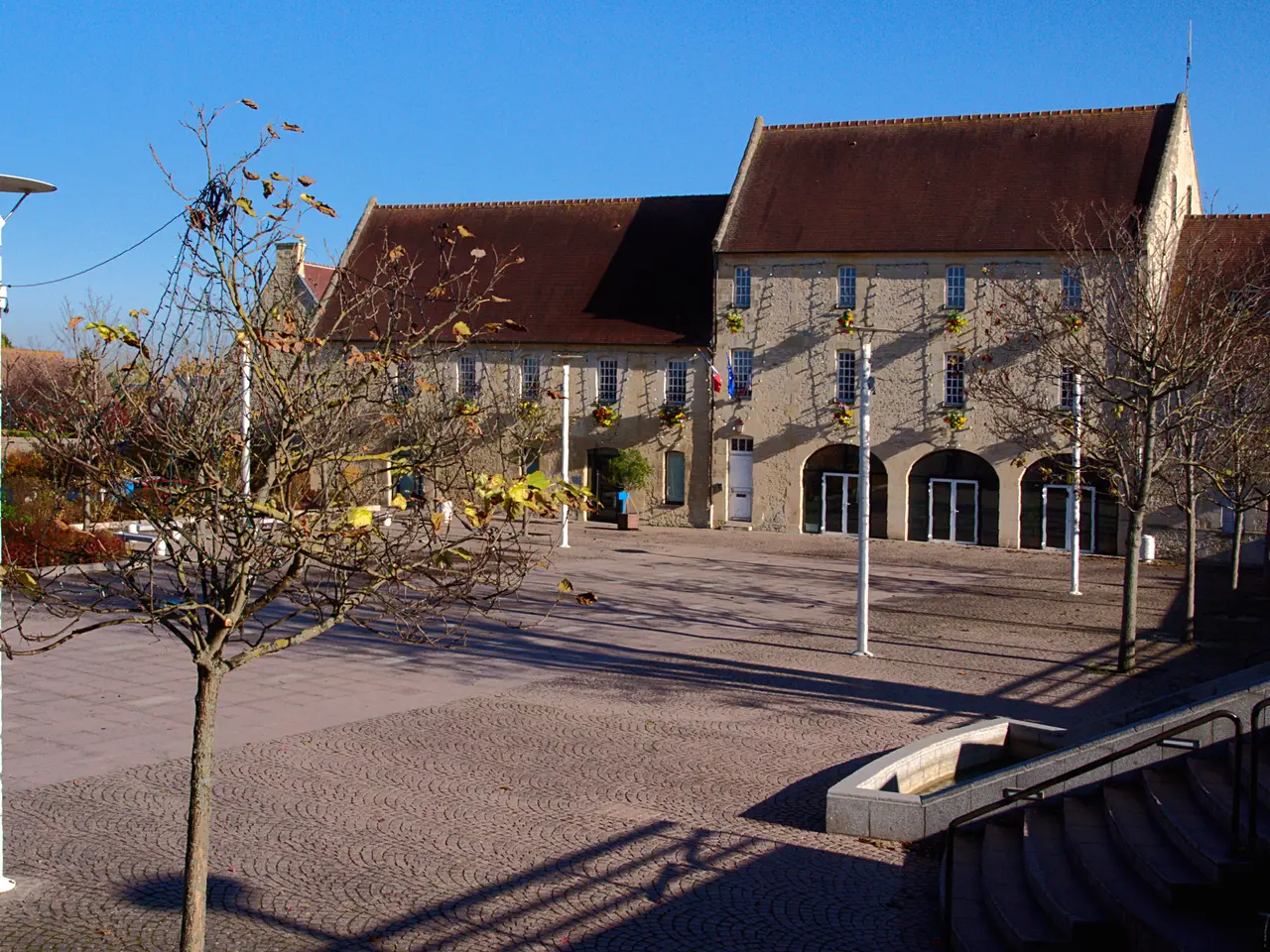 La mairie d’Ifs, ses bâtiments de pierre aux arcades et l’esplanade pavée qui s’étend devant, sous un ciel d’automne