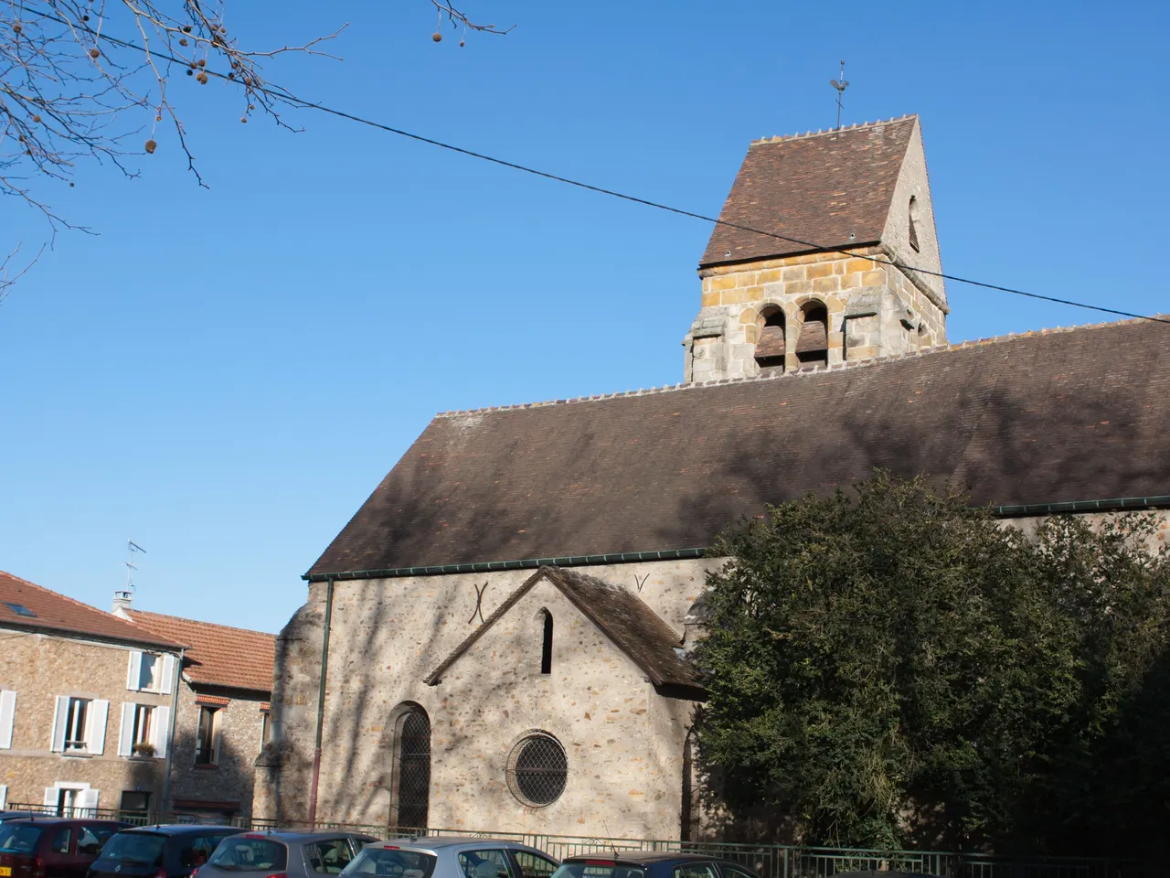 L’église Saint-Pierre d’Igny, ses murs de meulière et son clocher de pierre aux baies géminées, sous un ciel bleu, au-dessus de voitures garées.