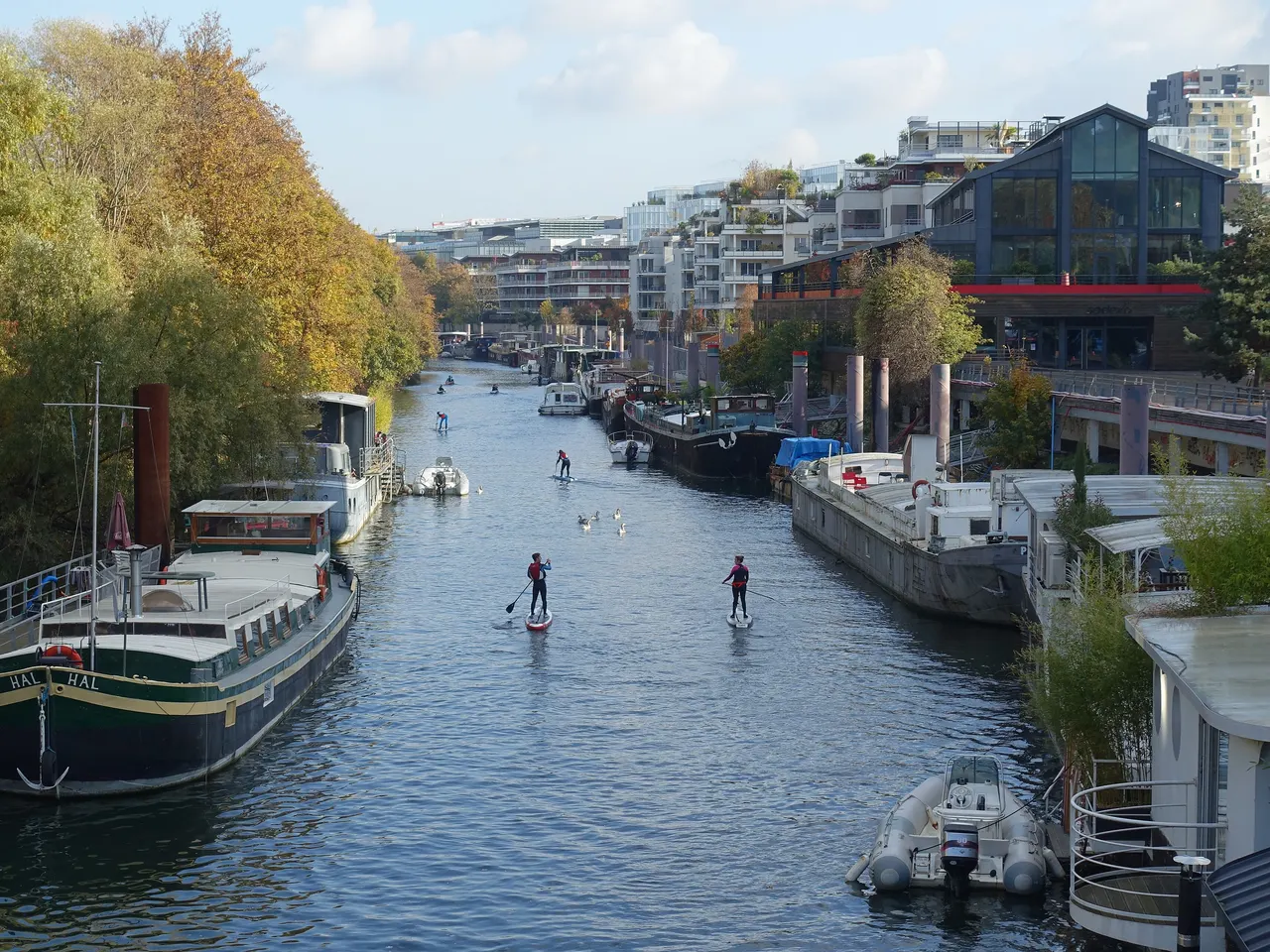 Le petit bras de Seine à Issy-les-Moulineaux, bordé de péniches, d’arbres d’automne et d’immeubles neufs, avec quelques pratiquants de paddle au loin.