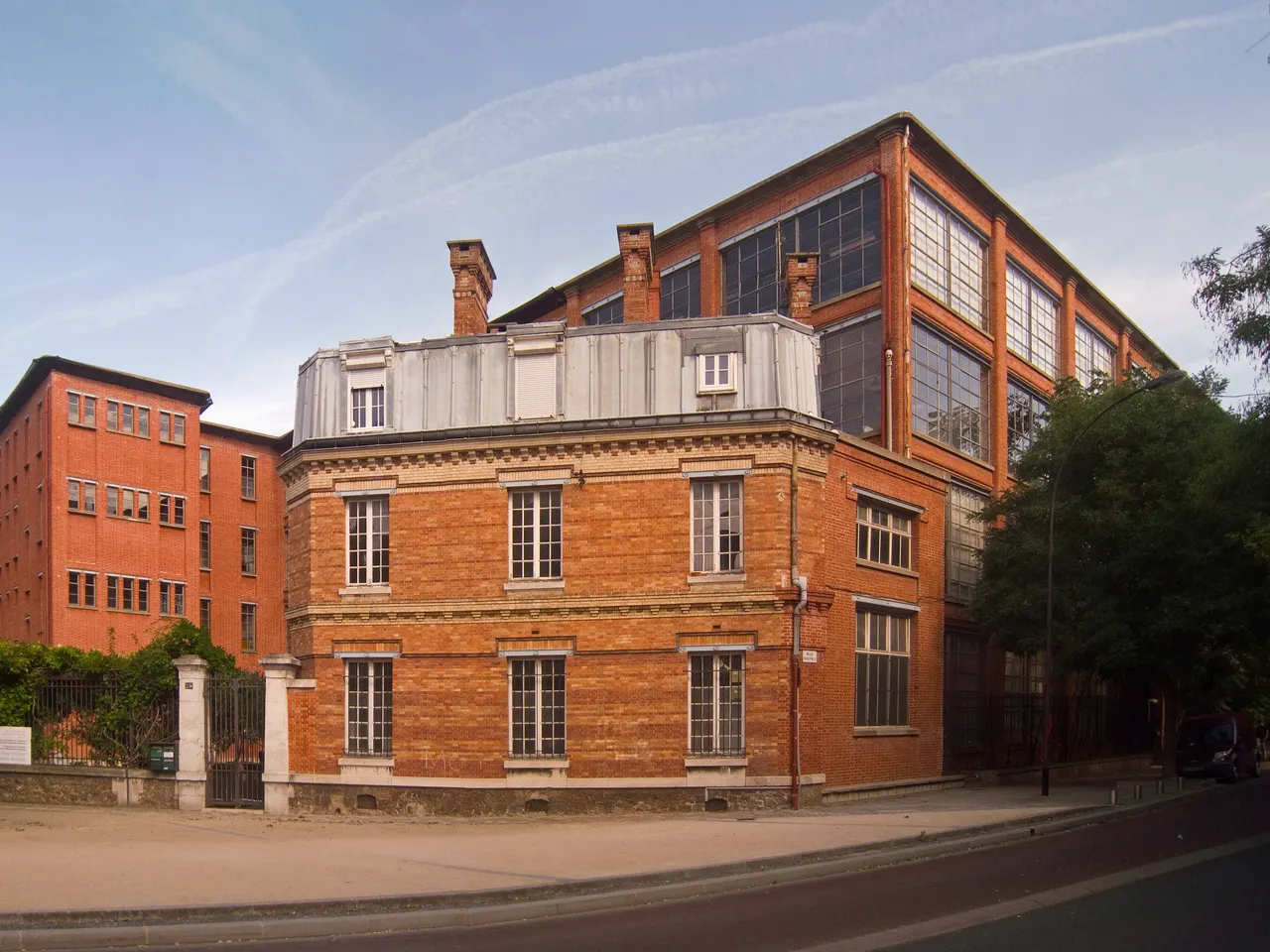 Les bâtiments de brique rouge de la Manufacture des œillets à Ivry-sur-Seine : une maison au toit de zinc au premier plan, un grand atelier aux larges verrières derrière.
