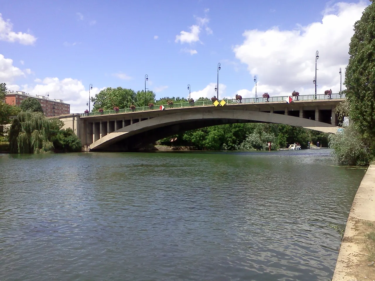 Le pont de Joinville, longue arche de béton au-dessus de la Marne, entre des saules et des rives boisées, sous un ciel bleu nuageux