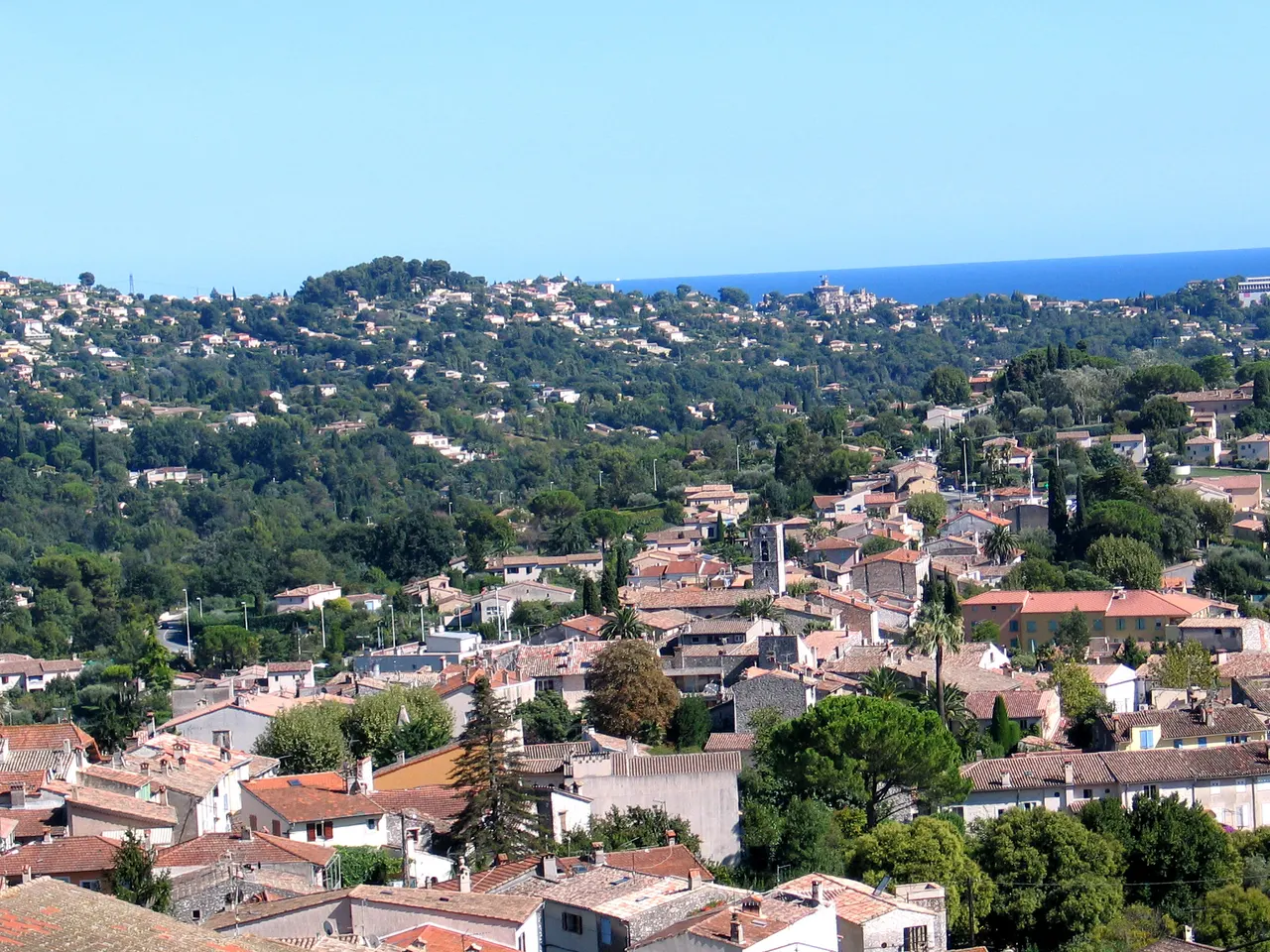 Les toits de tuiles de La Colle-sur-Loup et un clocher de pierre entre pins et palmiers, devant des collines boisées semées de villas, avec la mer à l’horizon.